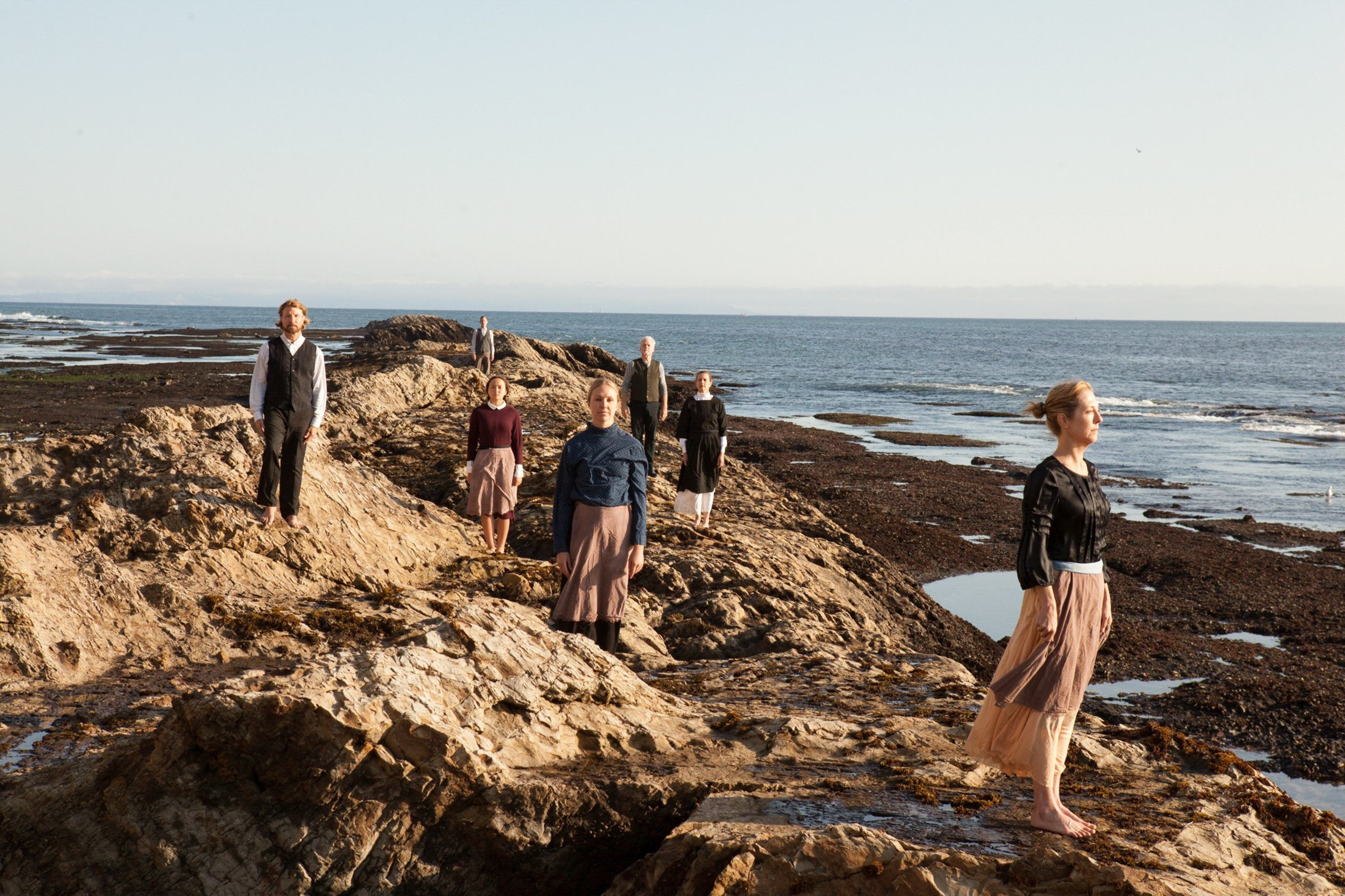 Six people stand spaced apart on rocky terrain by the sea, facing different directions, wearing modest, vintage-style clothing in neutral tones.