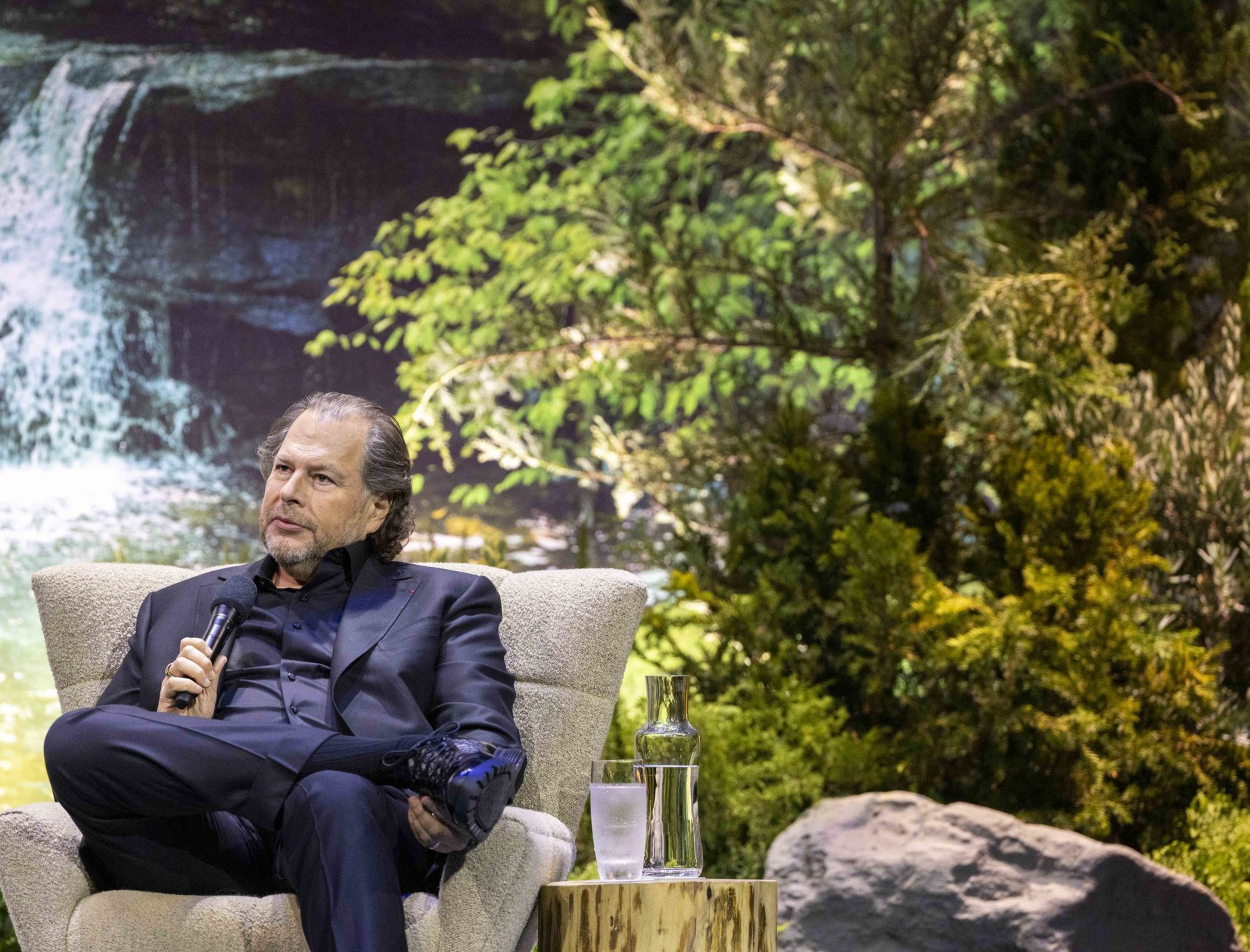 A man in a black suit sits in a chair, holding a microphone, with a lush, green forest and a waterfall backdrop behind him. A table with water and a glass is beside him.
