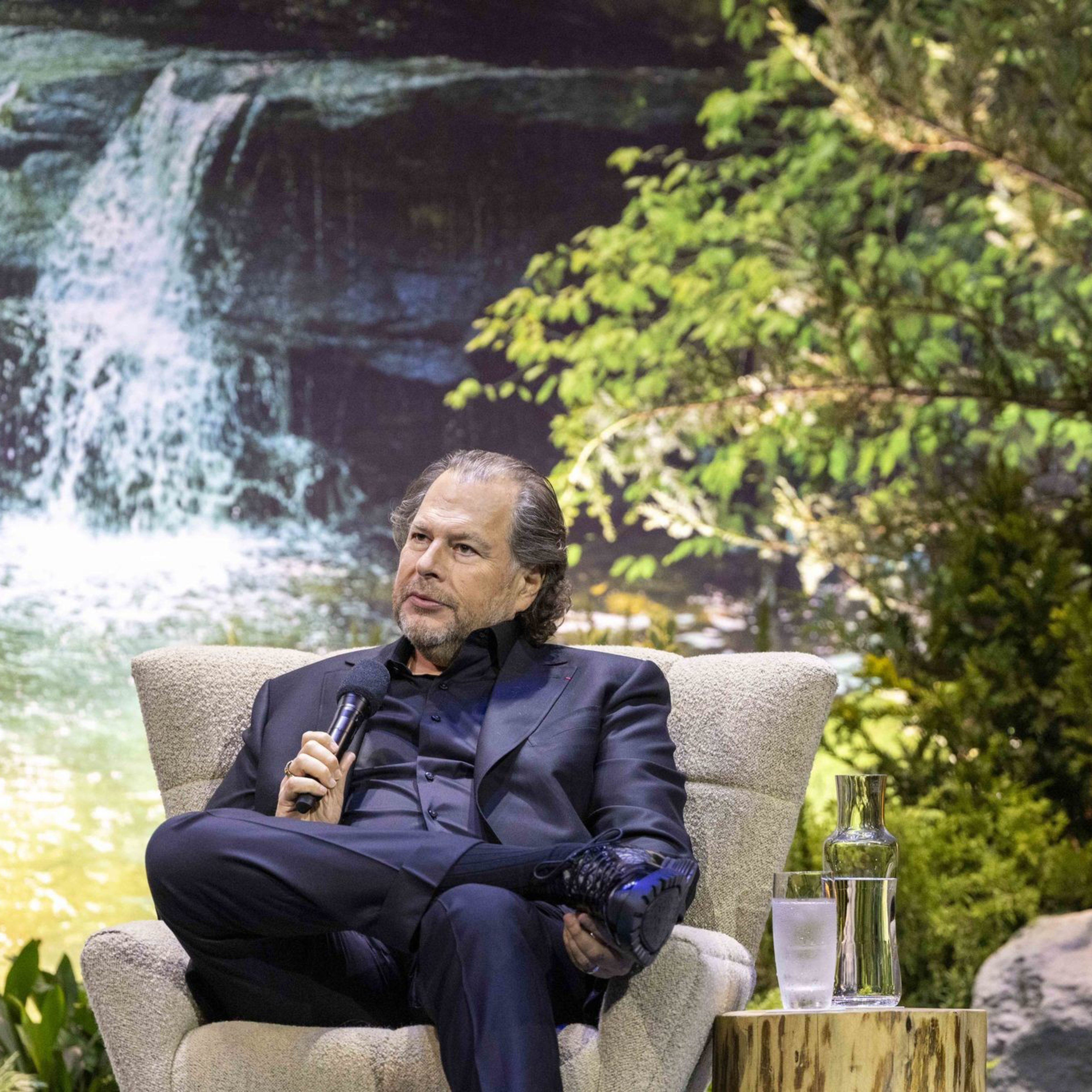 A man in a black suit sits in a chair, holding a microphone, with a lush, green forest and a waterfall backdrop behind him. A table with water and a glass is beside him.