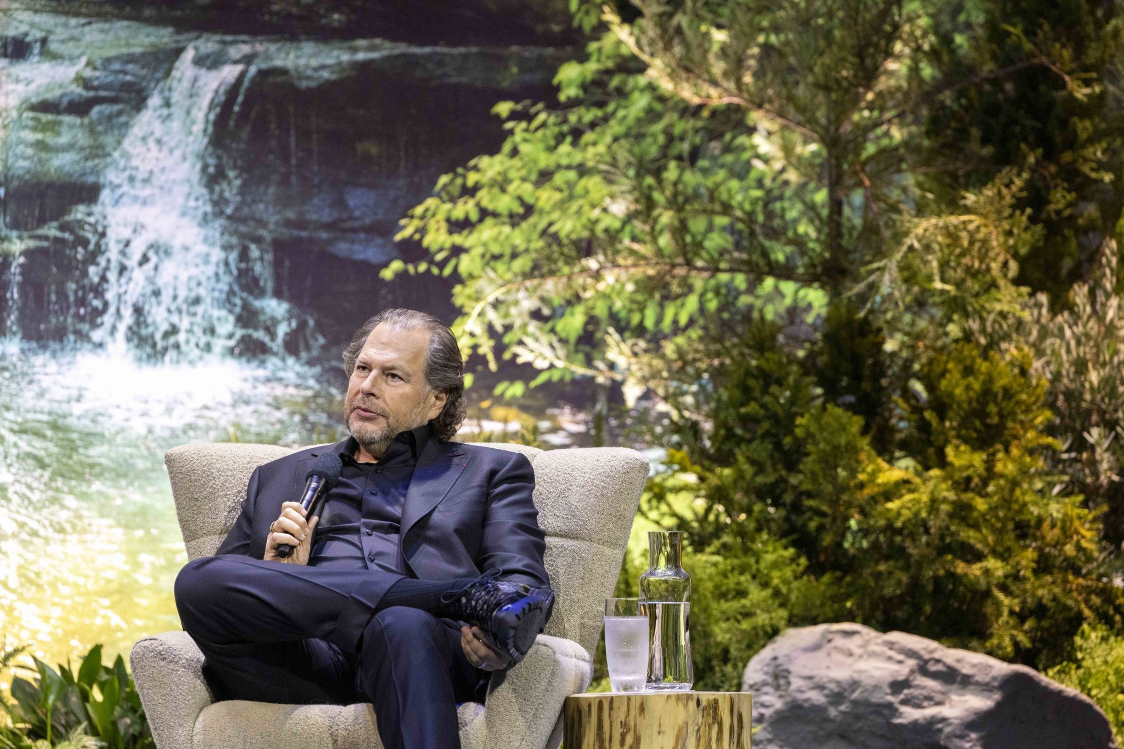 A man in a black suit sits in a chair, holding a microphone, with a lush, green forest and a waterfall backdrop behind him. A table with water and a glass is beside him.