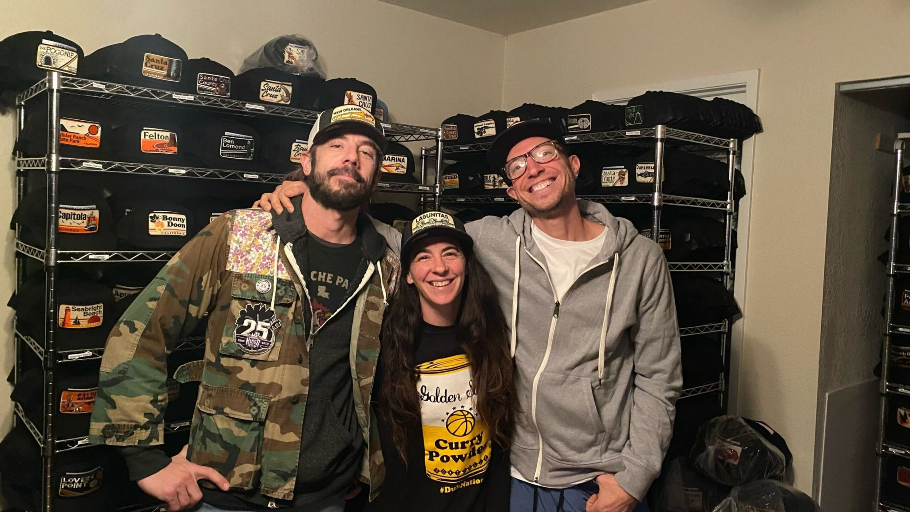 Three people wearing hats smile in front of shelves filled with neatly stacked folded black shirts featuring various colored patches.