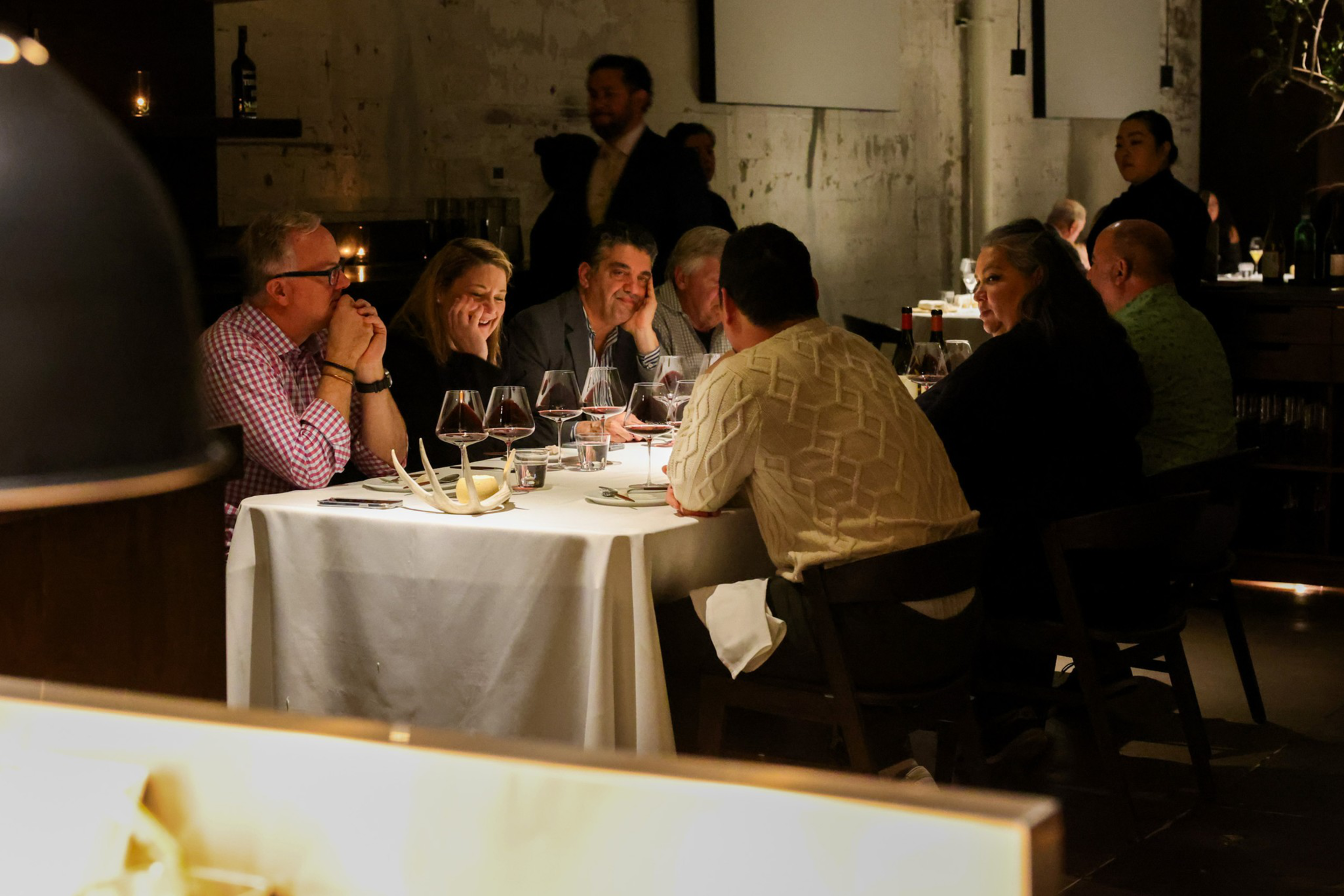 A group of people sit around a candlelit table in a dimly lit restaurant, engaging in conversation. Wine glasses and bottles are placed on the table.