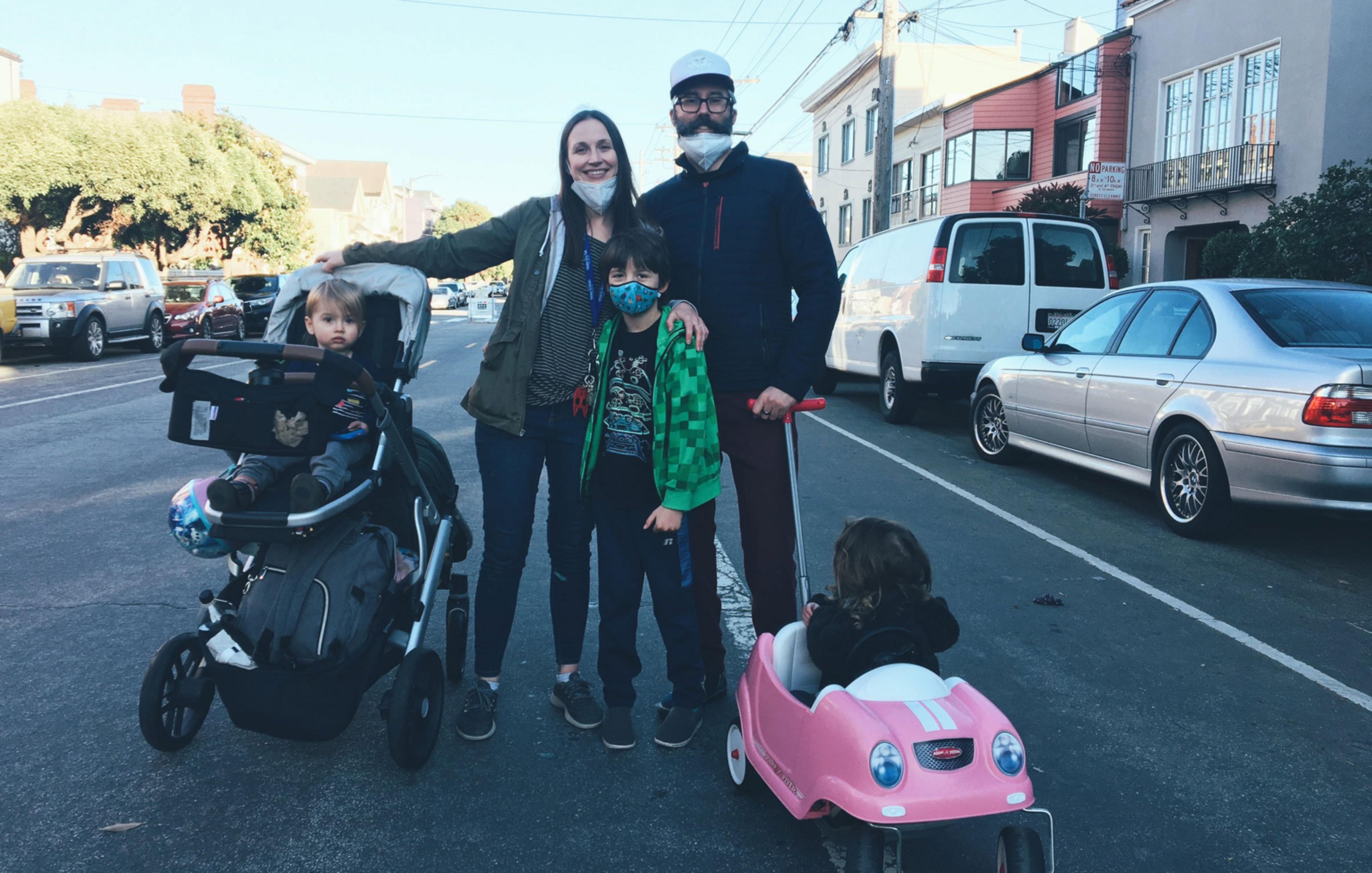 A family stands on a city street; a man and woman with two kids, one in a stroller and another sitting in a small pink toy car.