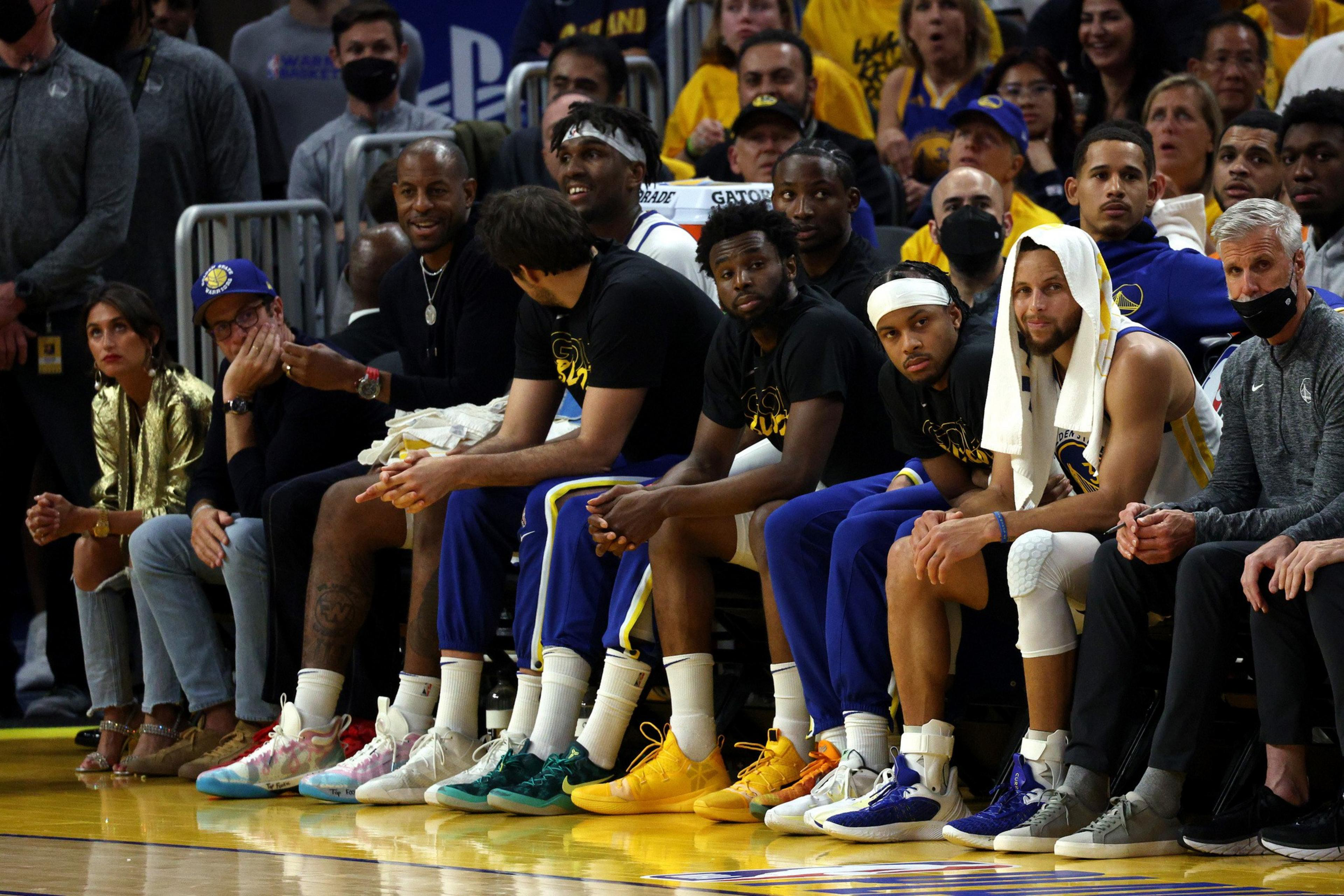 A group of people sit on a basketball bench, some in team jerseys and warm-ups, others in casual clothing. They appear attentive, with a towel seen on one player's head.