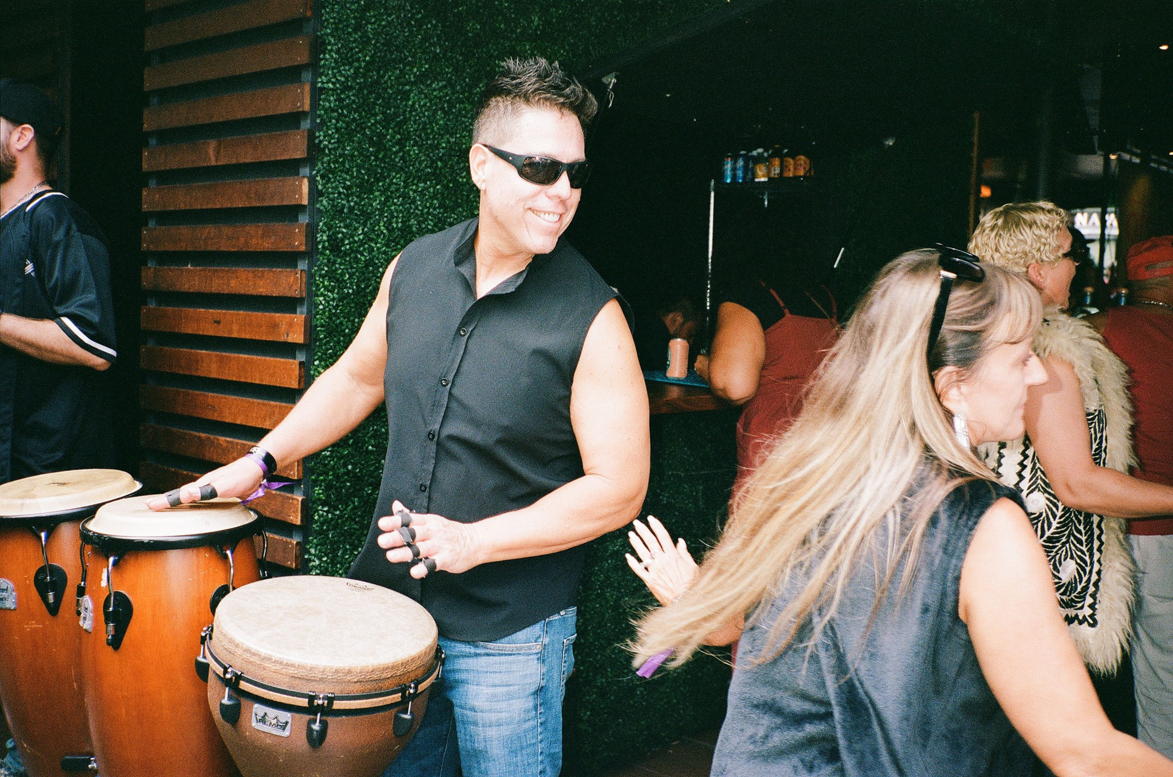 A man wearing sunglasses and a sleeveless black shirt plays conga drums while smiling, as two women nearby move energetically.