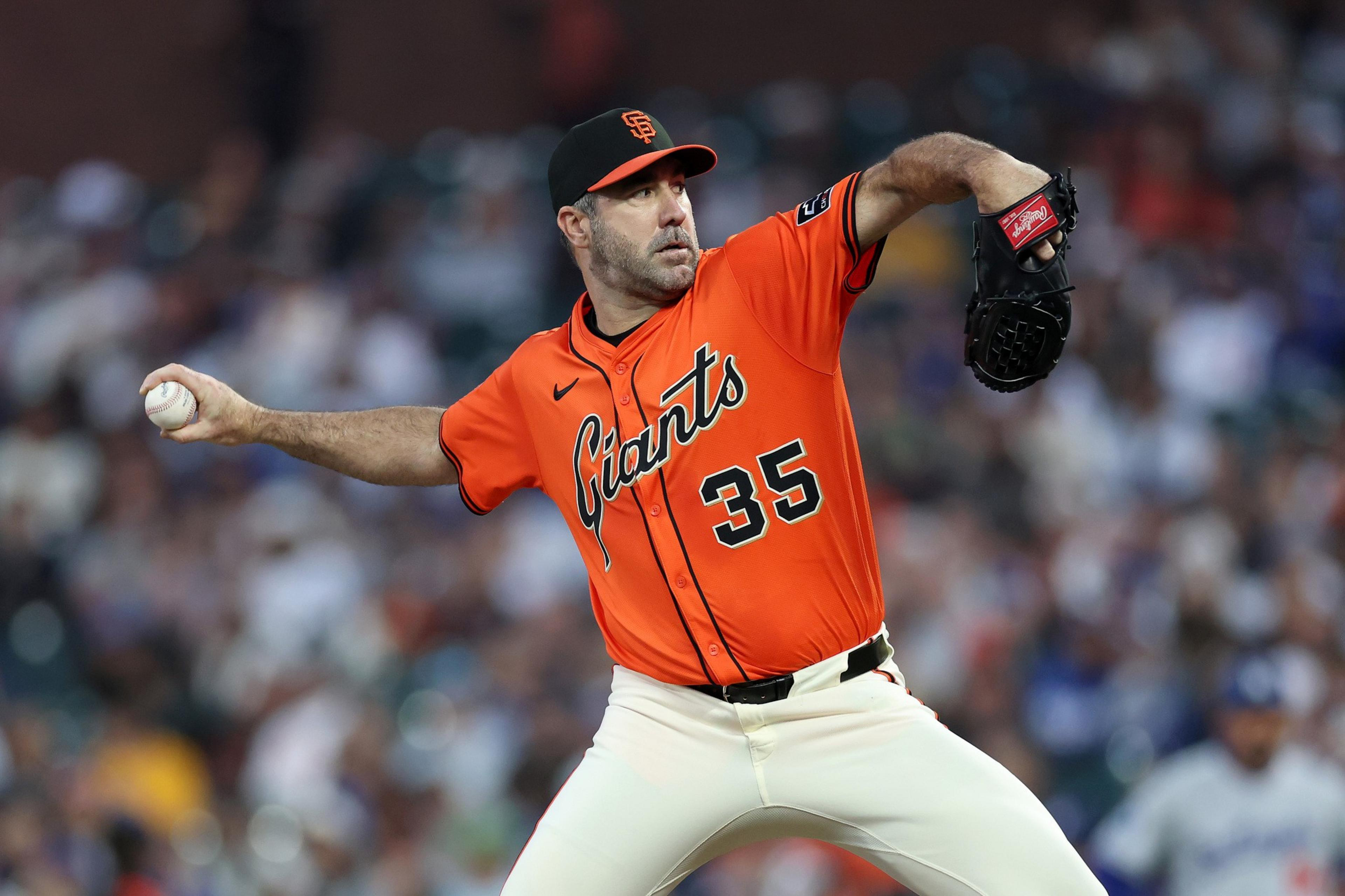 A baseball pitcher in an orange Giants jersey, number 35, is in mid-pitch with a focused expression during a game in front of a blurred crowd.