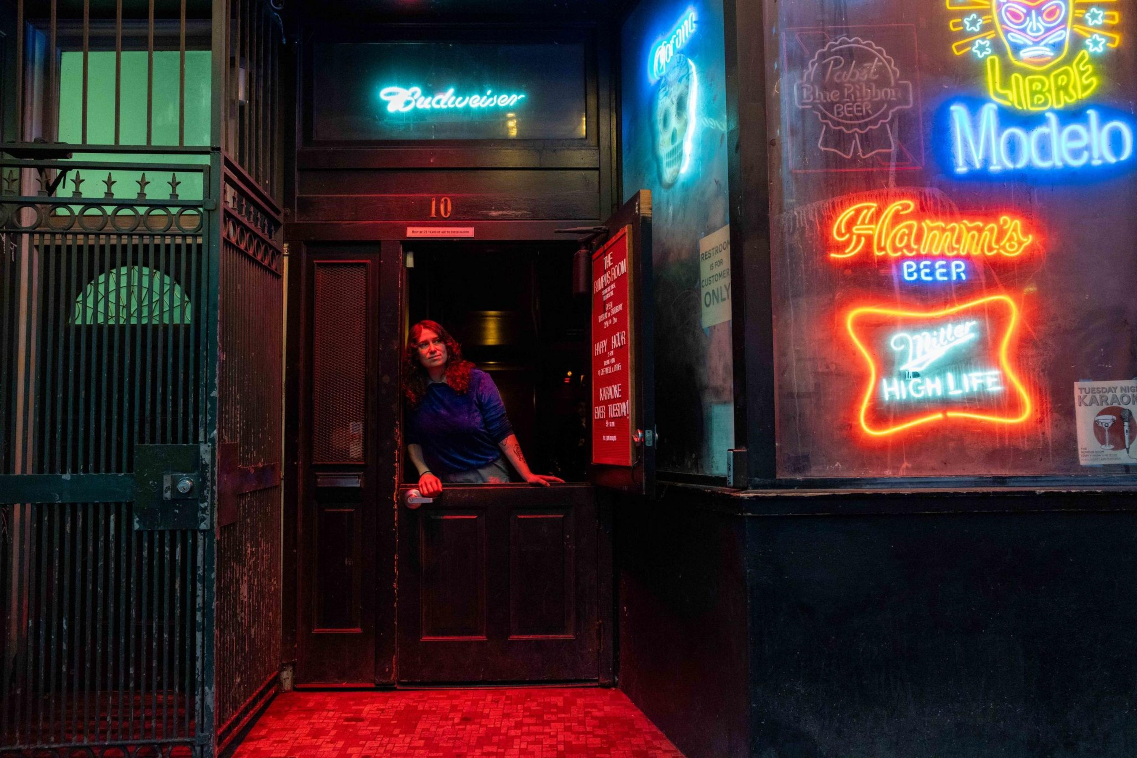 A person stands in a dimly lit doorway, surrounded by colorful neon beer signs. The atmosphere appears vibrant and inviting.