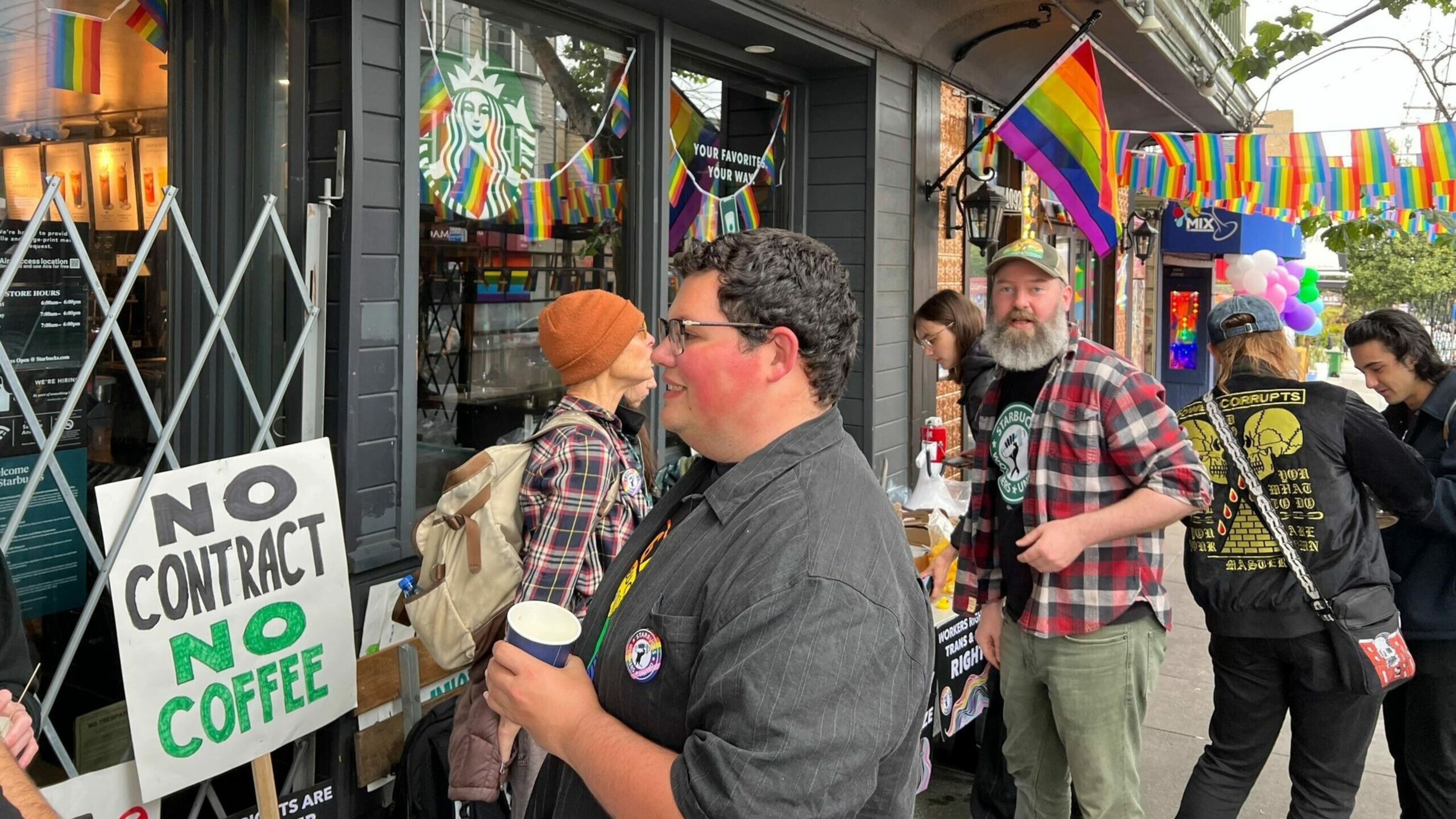 People gather outside a Starbucks adorned with Pride flags, one holding a sign that reads "NO CONTRACT NO COFFEE," likely protesting for workers' rights.