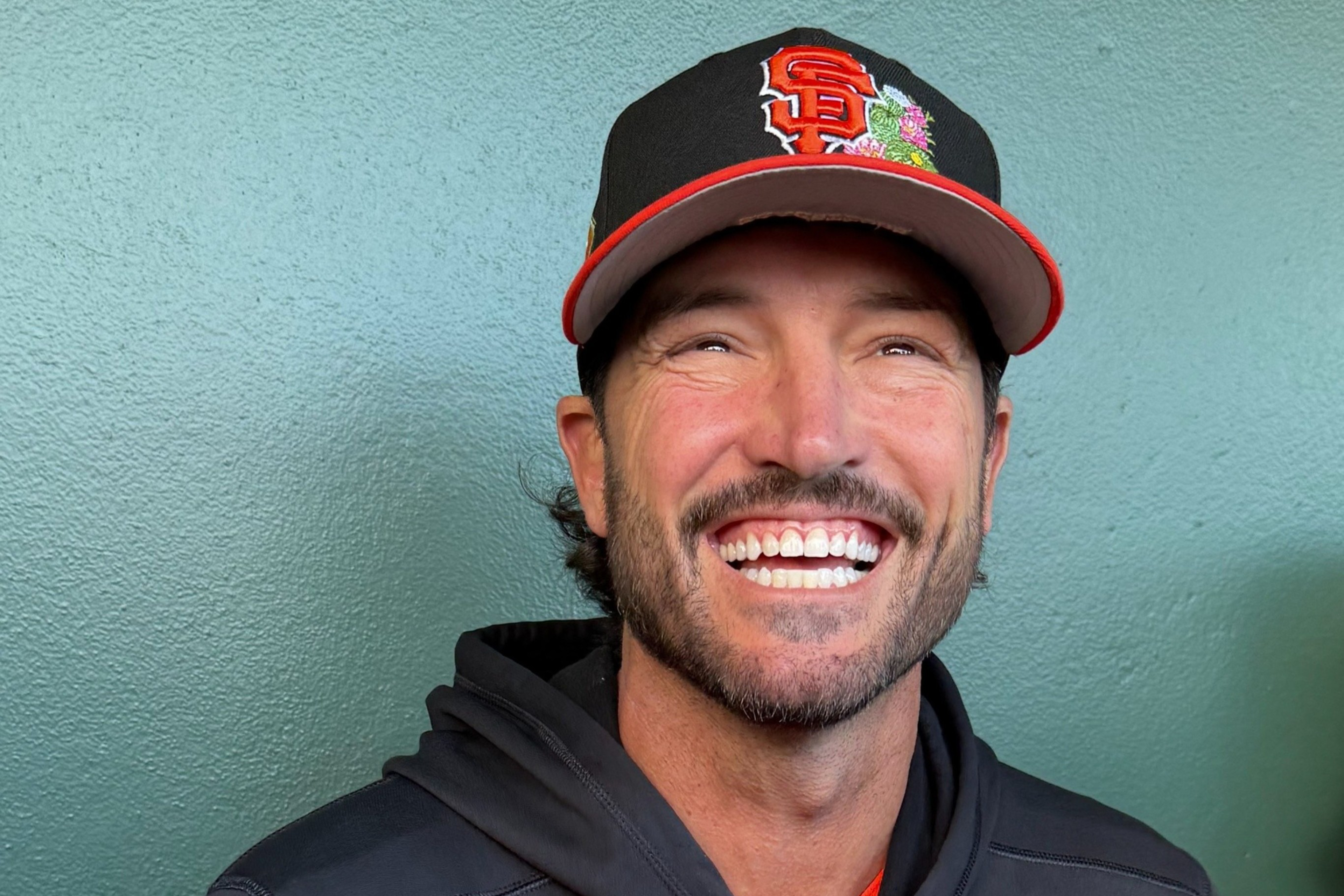 A man with a beard is smiling broadly, wearing a black and orange San Francisco Giants baseball cap and a black hoodie.