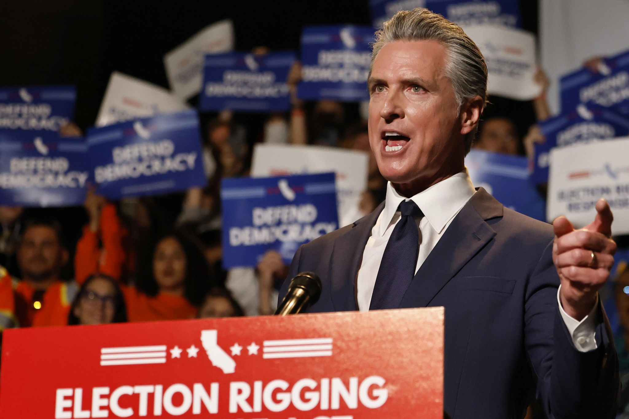A man in a suit speaks passionately at a podium labeled "Election Rigging Response Act," with a crowd holding "Defend Democracy" signs behind him.