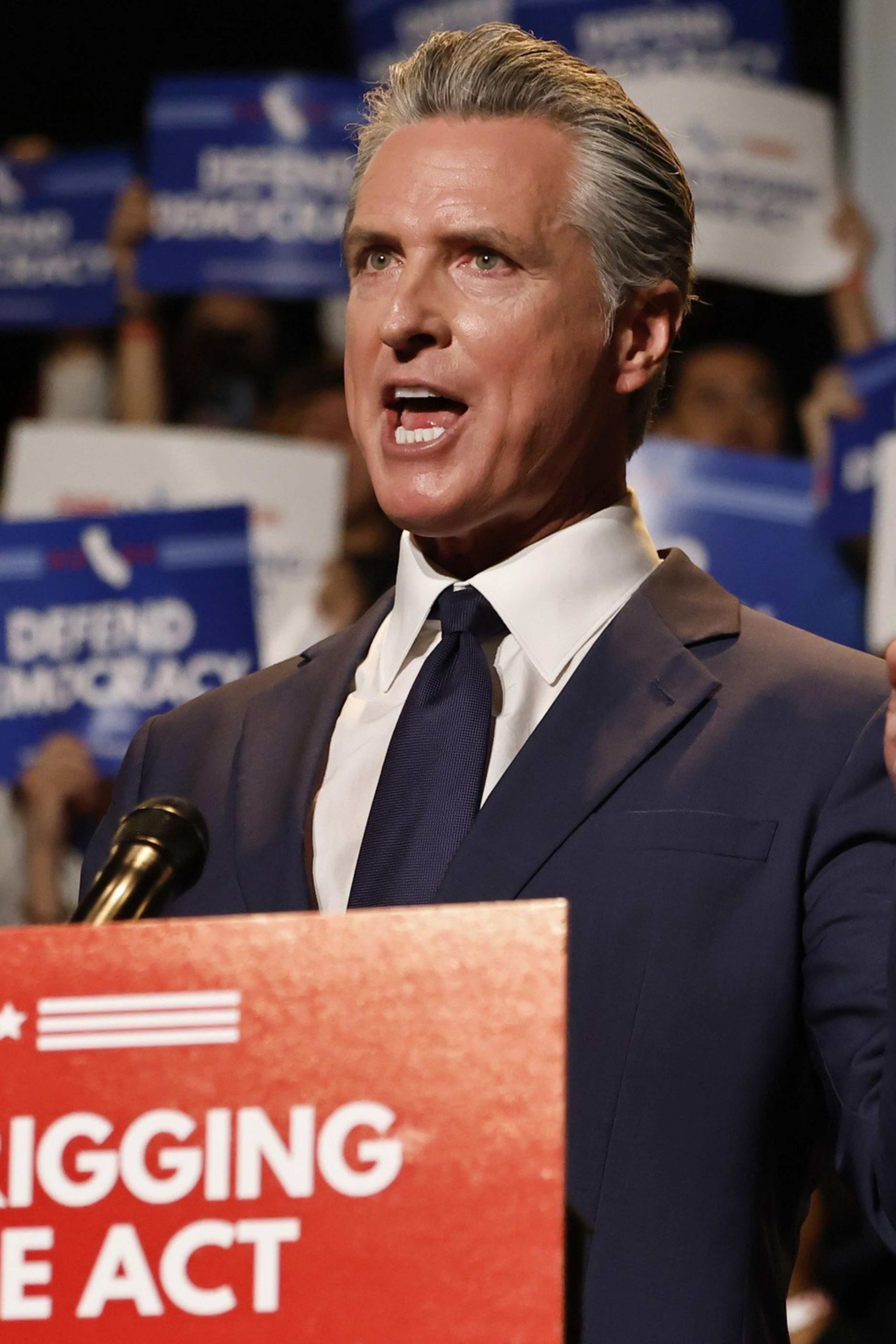 A man in a suit speaks passionately at a podium labeled "Election Rigging Response Act," with a crowd holding "Defend Democracy" signs behind him.