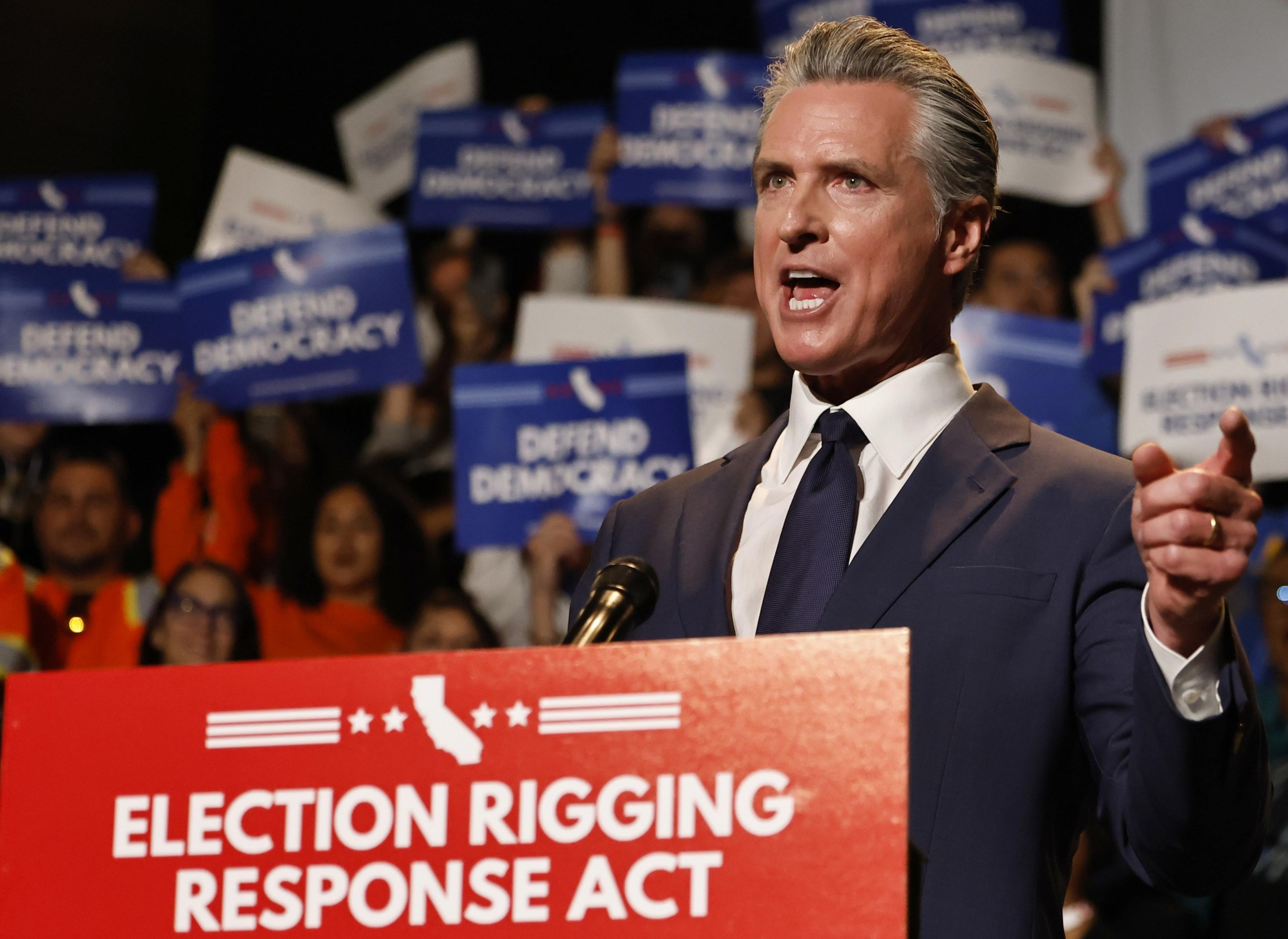 A man in a suit speaks passionately at a podium labeled "Election Rigging Response Act," with a crowd holding "Defend Democracy" signs behind him.