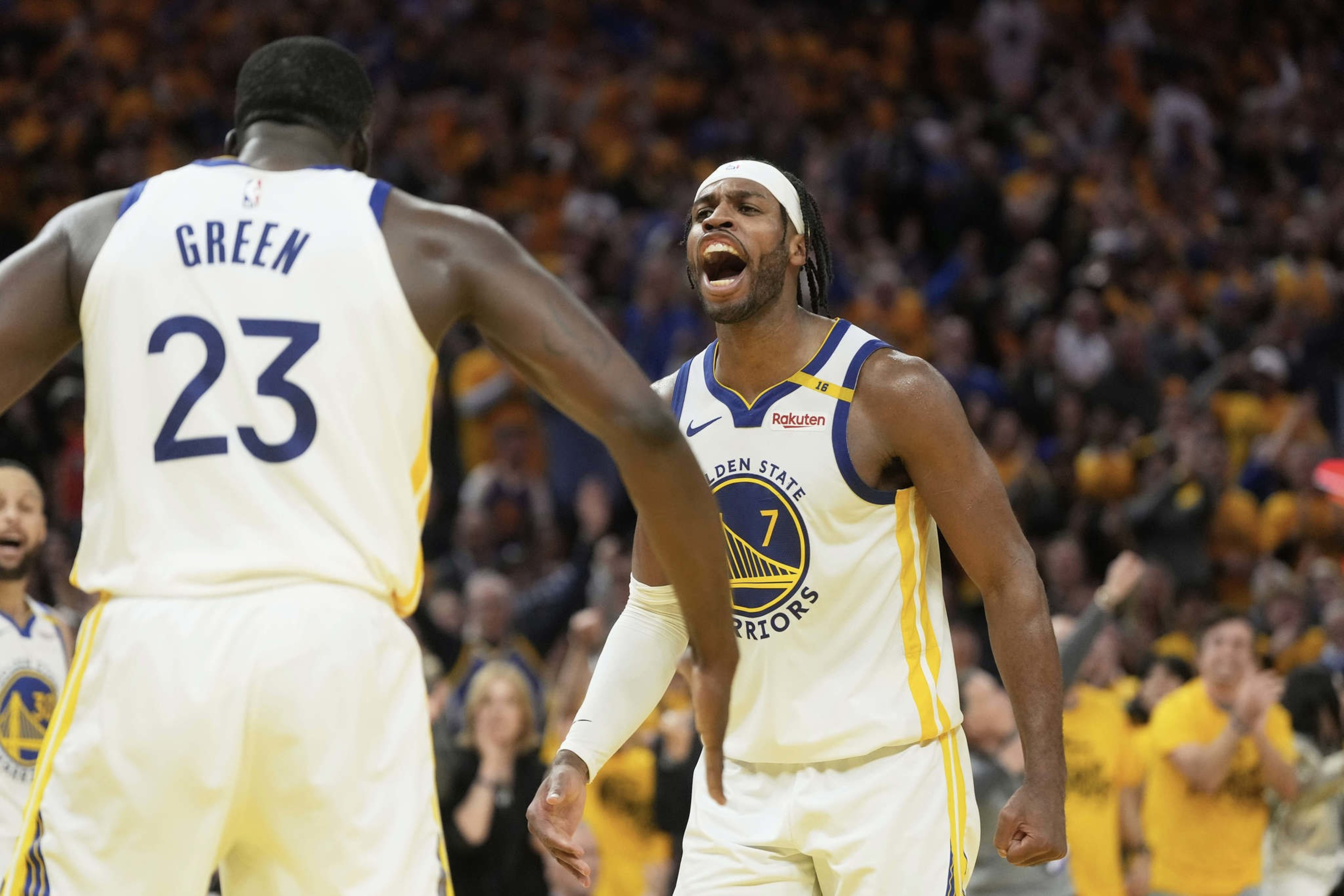 Two basketball players in Golden State Warriors jerseys are celebrating on the court. One, facing away, has "Green" on the jersey. The crowd in the background is cheering.