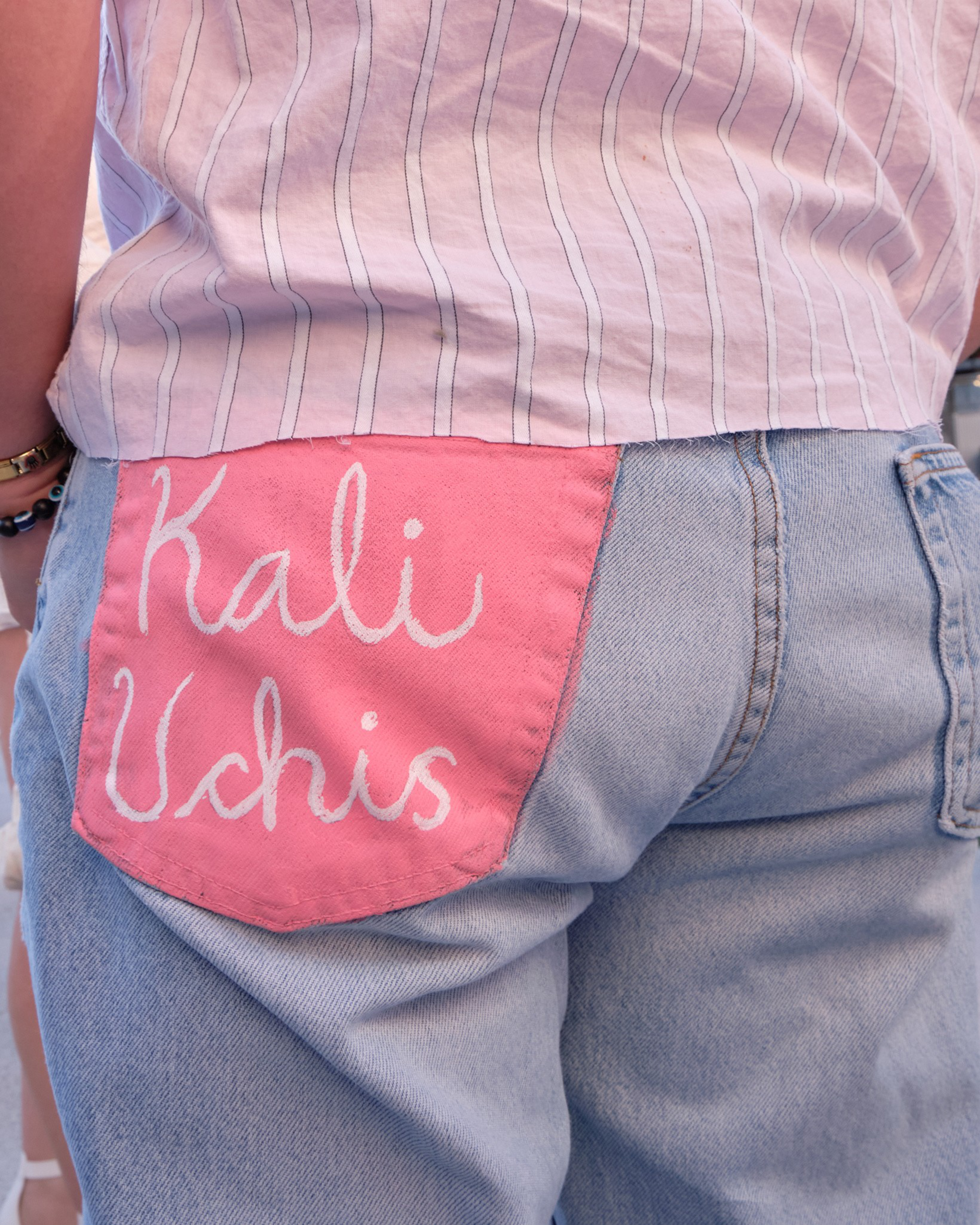 Light blue jeans feature a pink back pocket with the name "Kali Uchis" written in white cursive, paired with a light striped shirt.