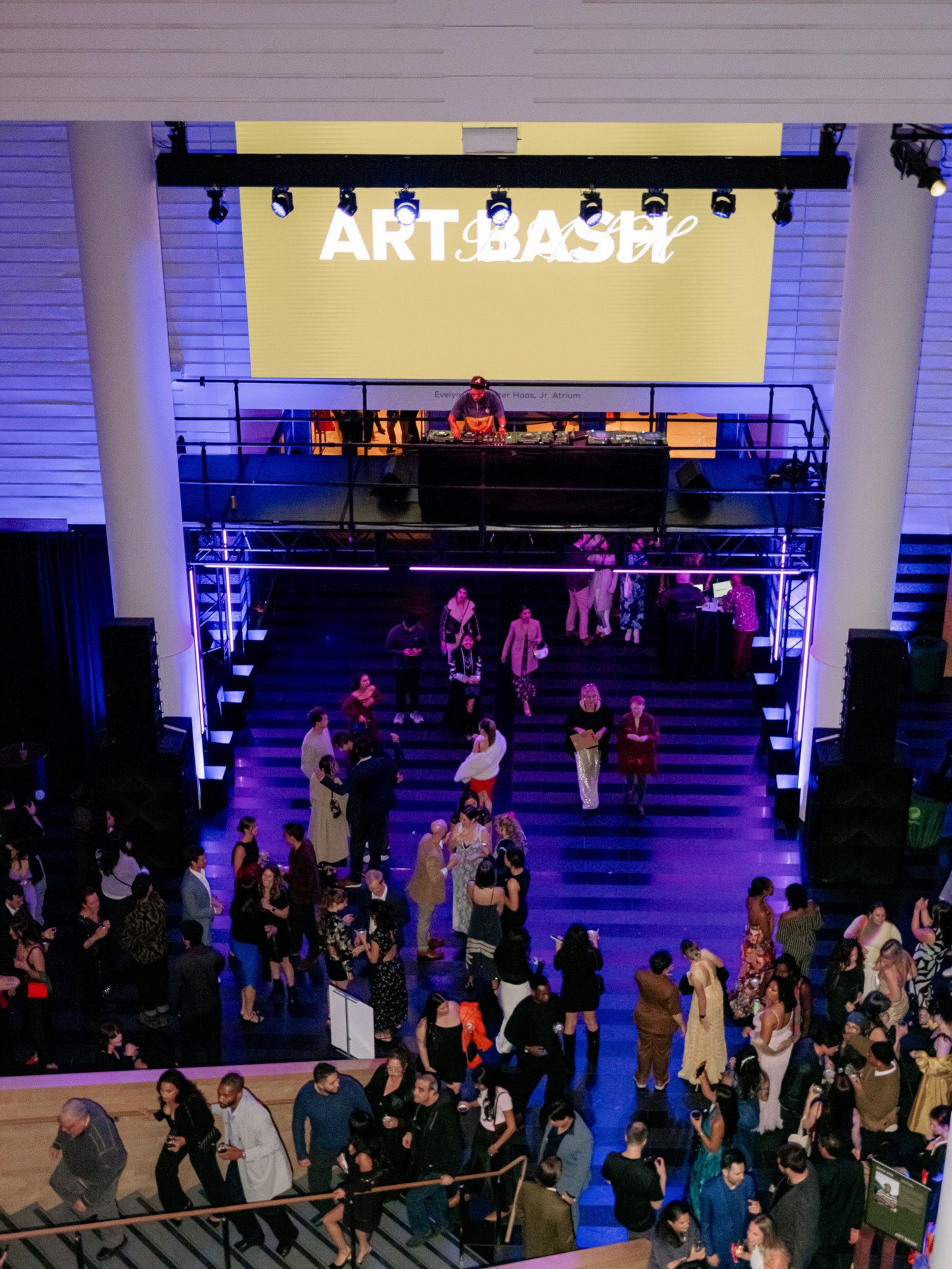 The image shows a lively event with a crowd gathered in a large venue. A DJ is performing on an elevated stage with the words &quot;ART BASH&quot; projected above.