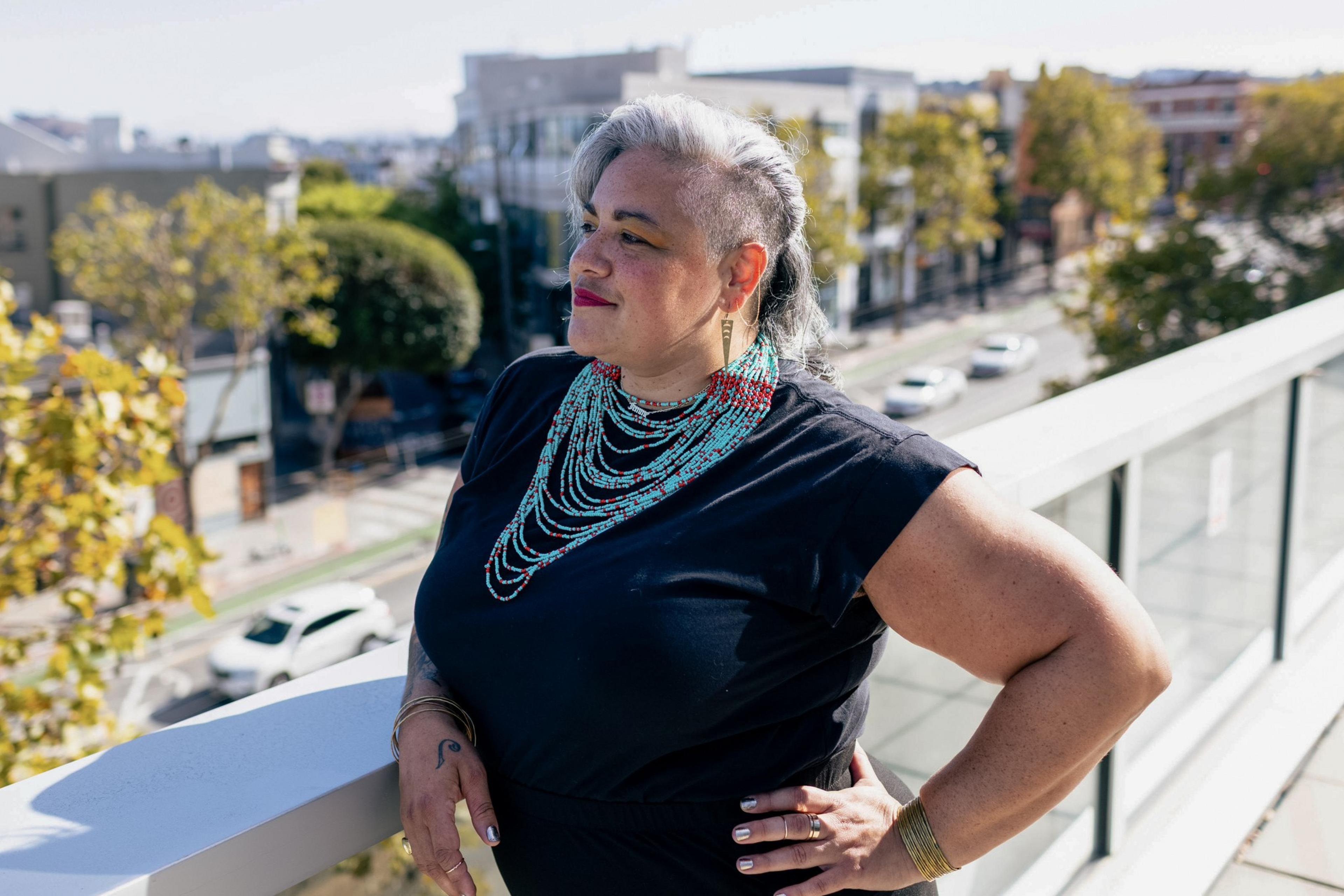 A woman with short gray and black hair stands on a balcony, wearing a black top and layered turquoise necklaces, looking confidently to the side.