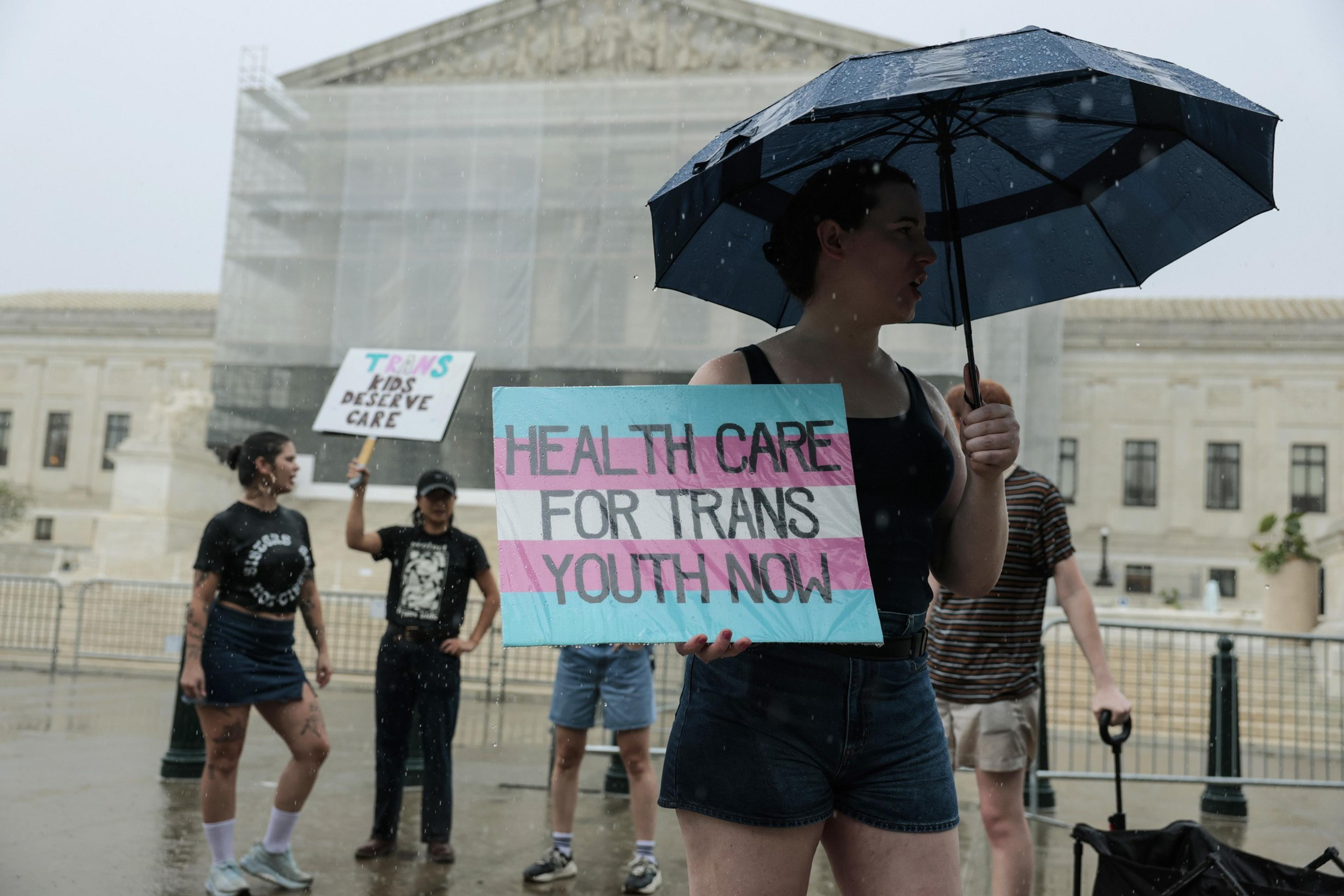 People are standing in the rain holding signs advocating for trans youth healthcare outside a large government building. One person holds an umbrella.