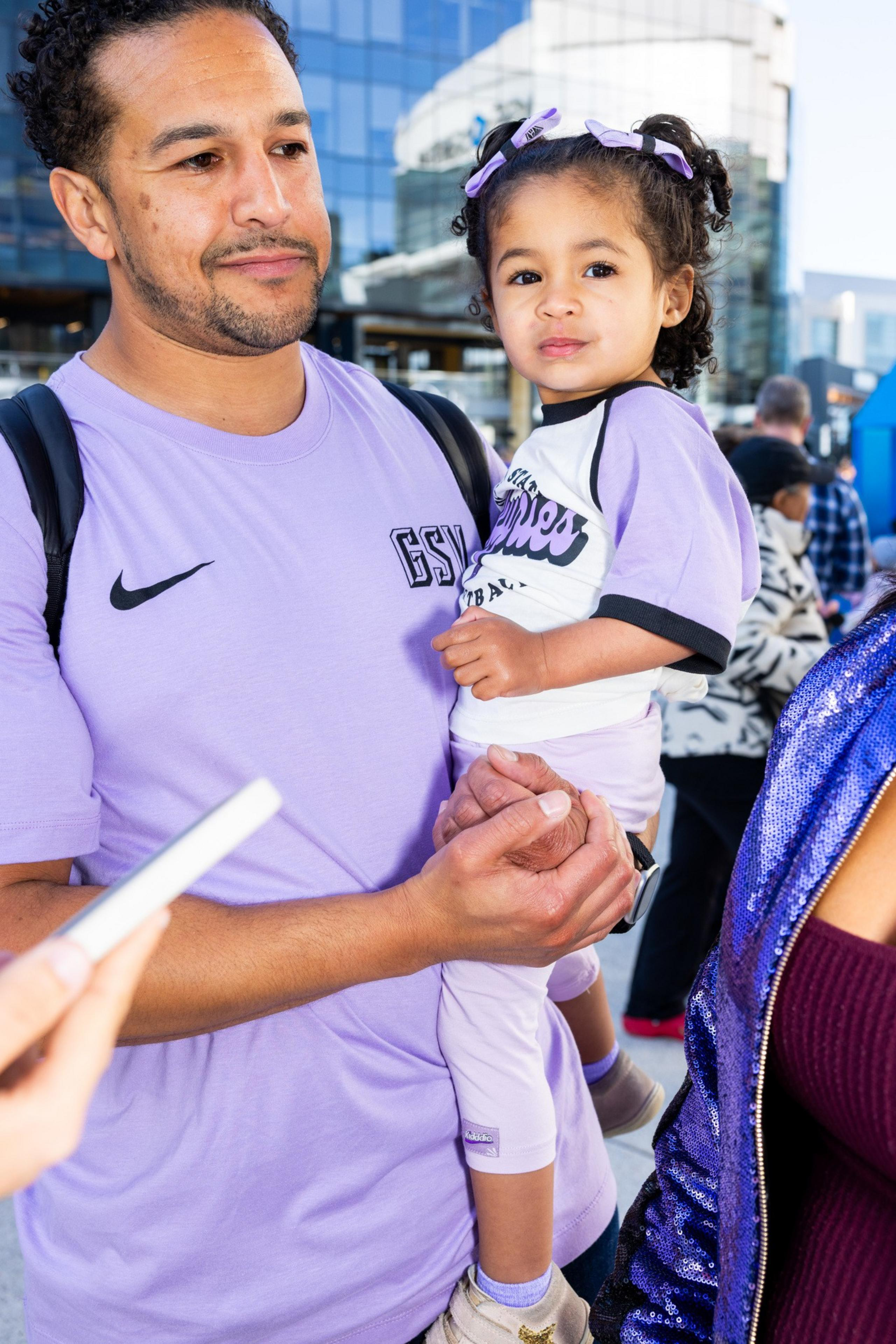A man in a purple shirt holds a young child wearing a purple and white outfit. They are outdoors, with a building and other people in the background.