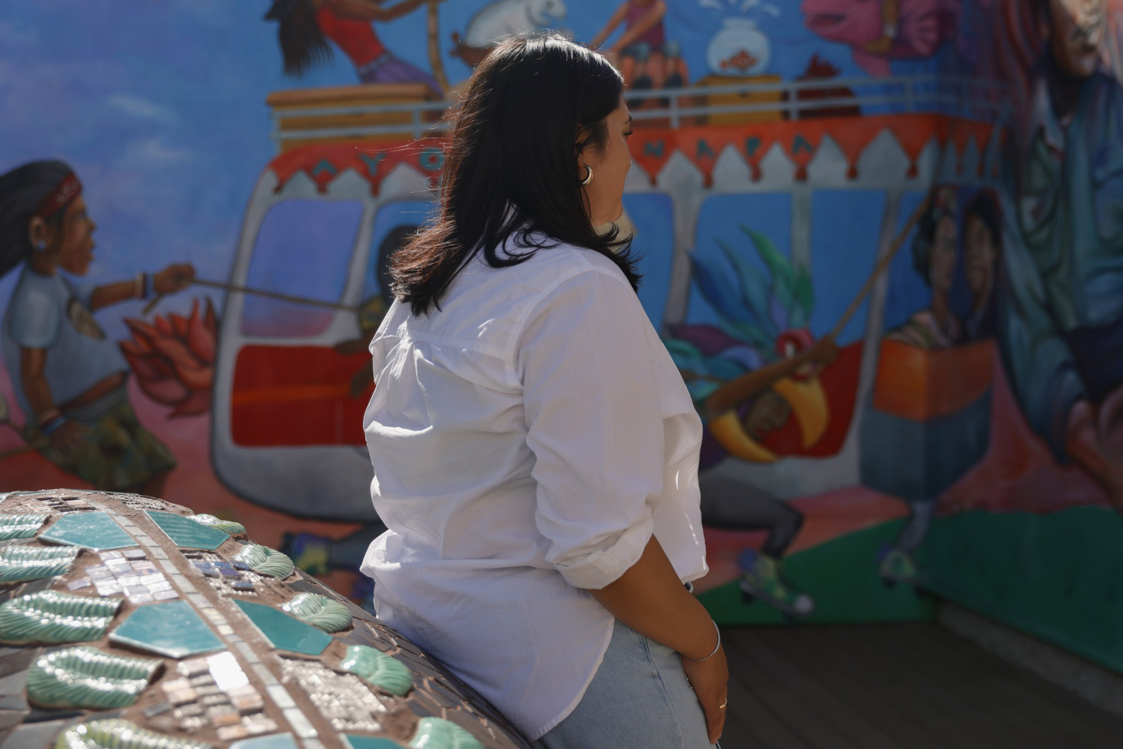 A woman with dark hair wearing a white shirt and jeans sits on a mosaic bench, facing a colorful mural with people in dynamic poses.