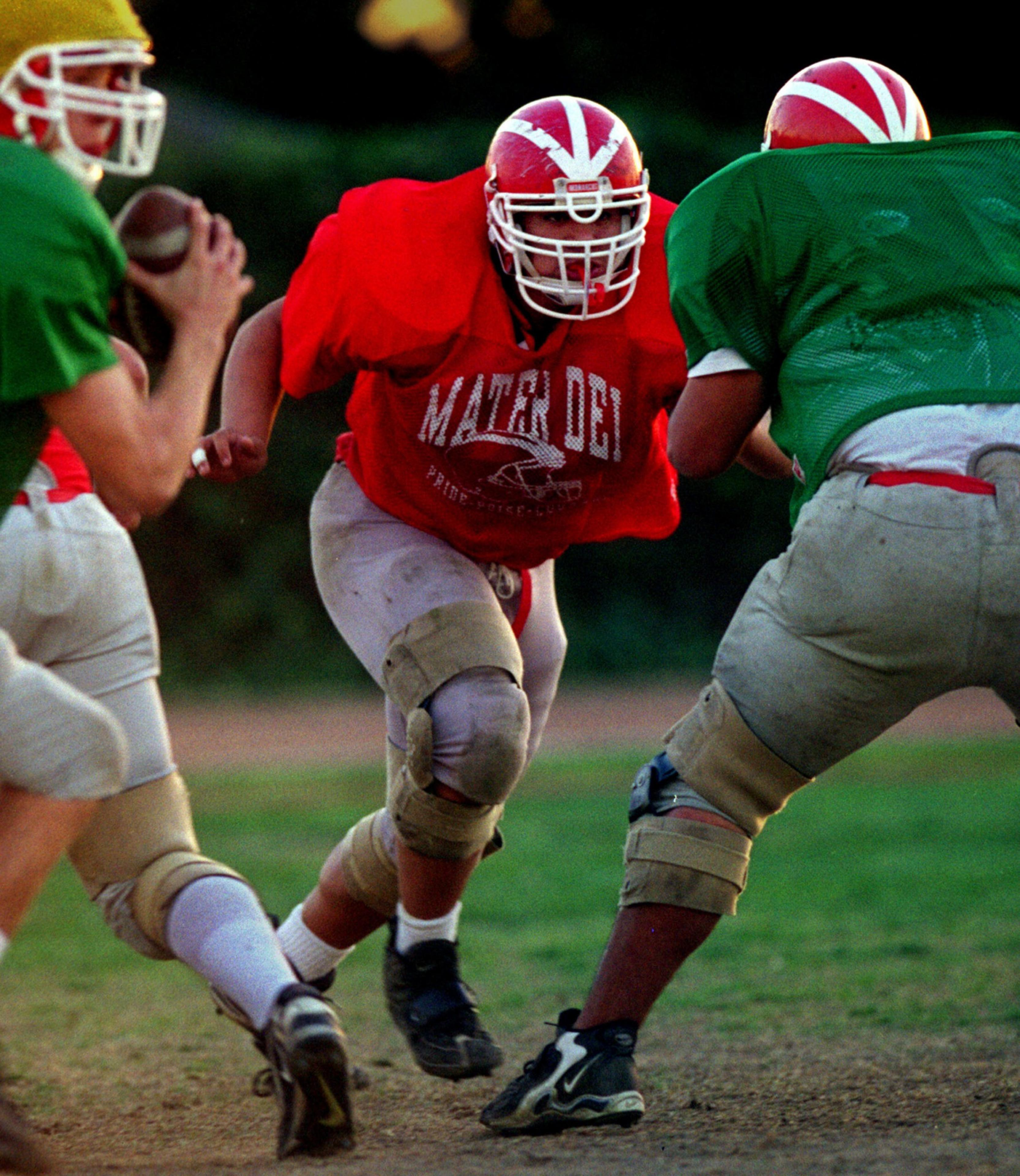 A football player in a red jersey and white helmet charges toward two opponents wearing green jerseys and helmets on a grassy field during practice.