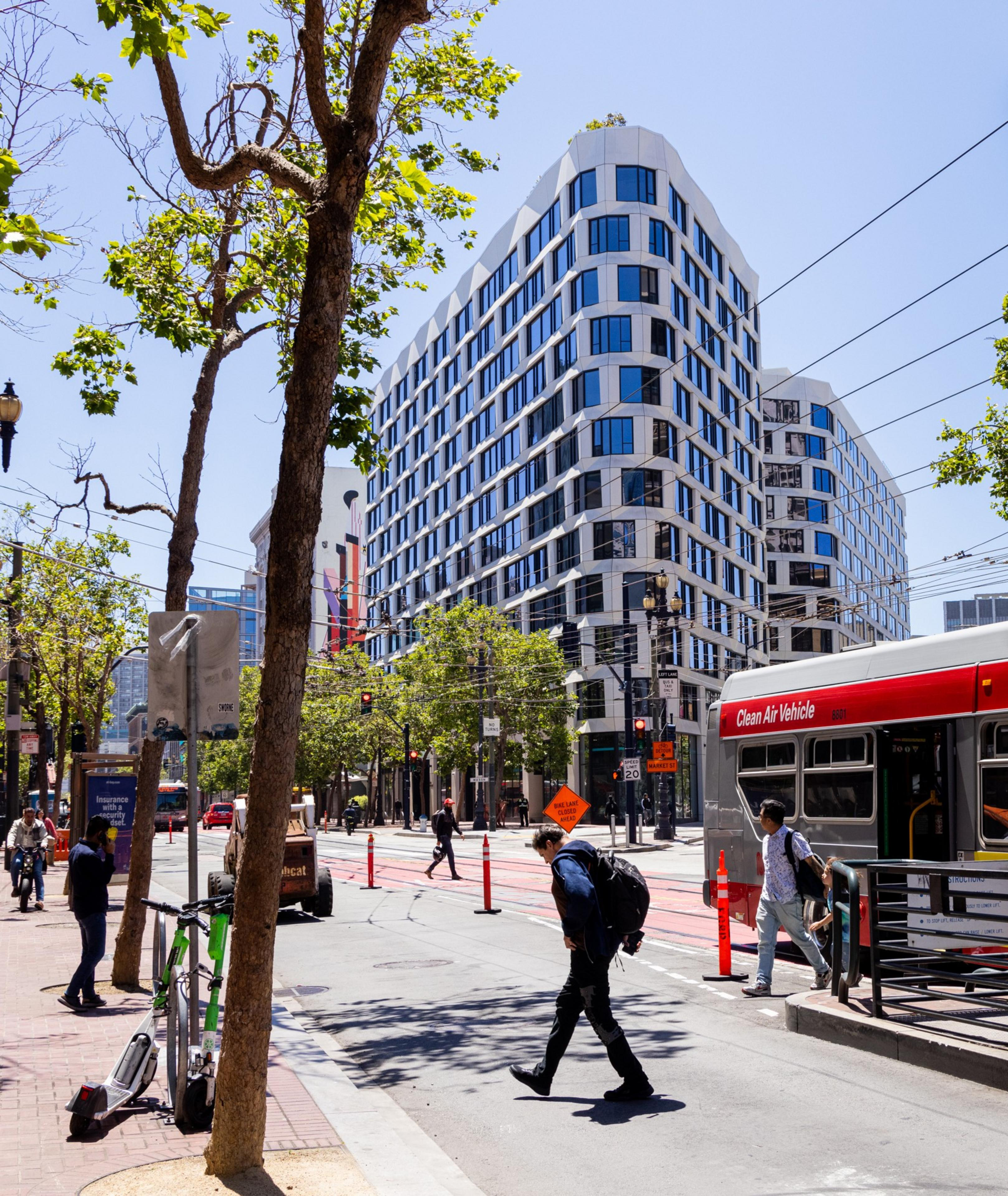 A busy urban street scene with people walking, a modern multi-story building, trees, a city bus labeled &quot;Clean Air Vehicle,&quot; and scattered orange traffic cones.