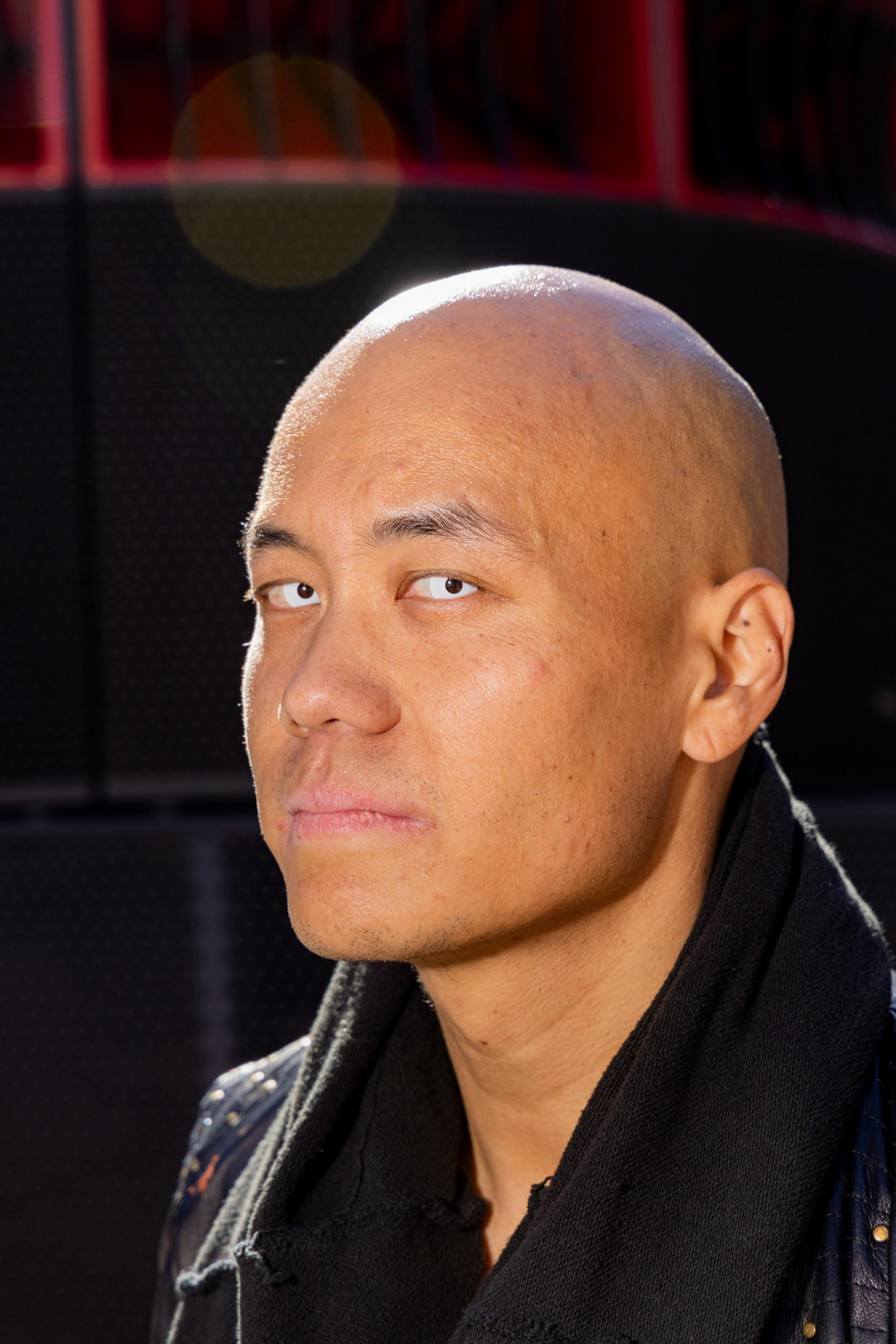 A bald man with a serious expression is looking slightly upward. He wears a dark jacket and scarf, and the background features dark, textured surfaces with red highlights.