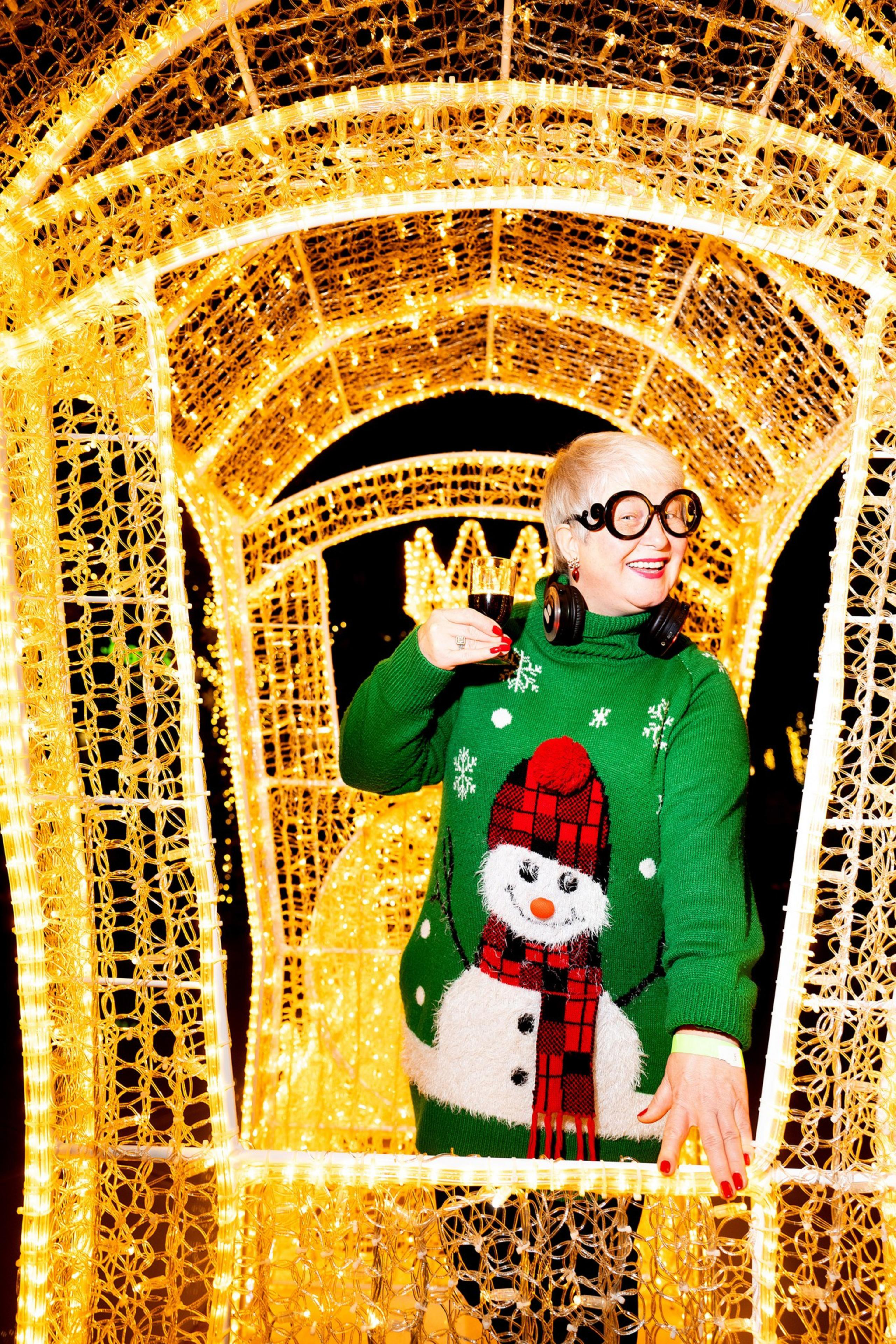 A person in a green sweater with a snowman design stands under a tunnel of golden lights, smiling and holding a glass, wearing large round glasses and headphones.