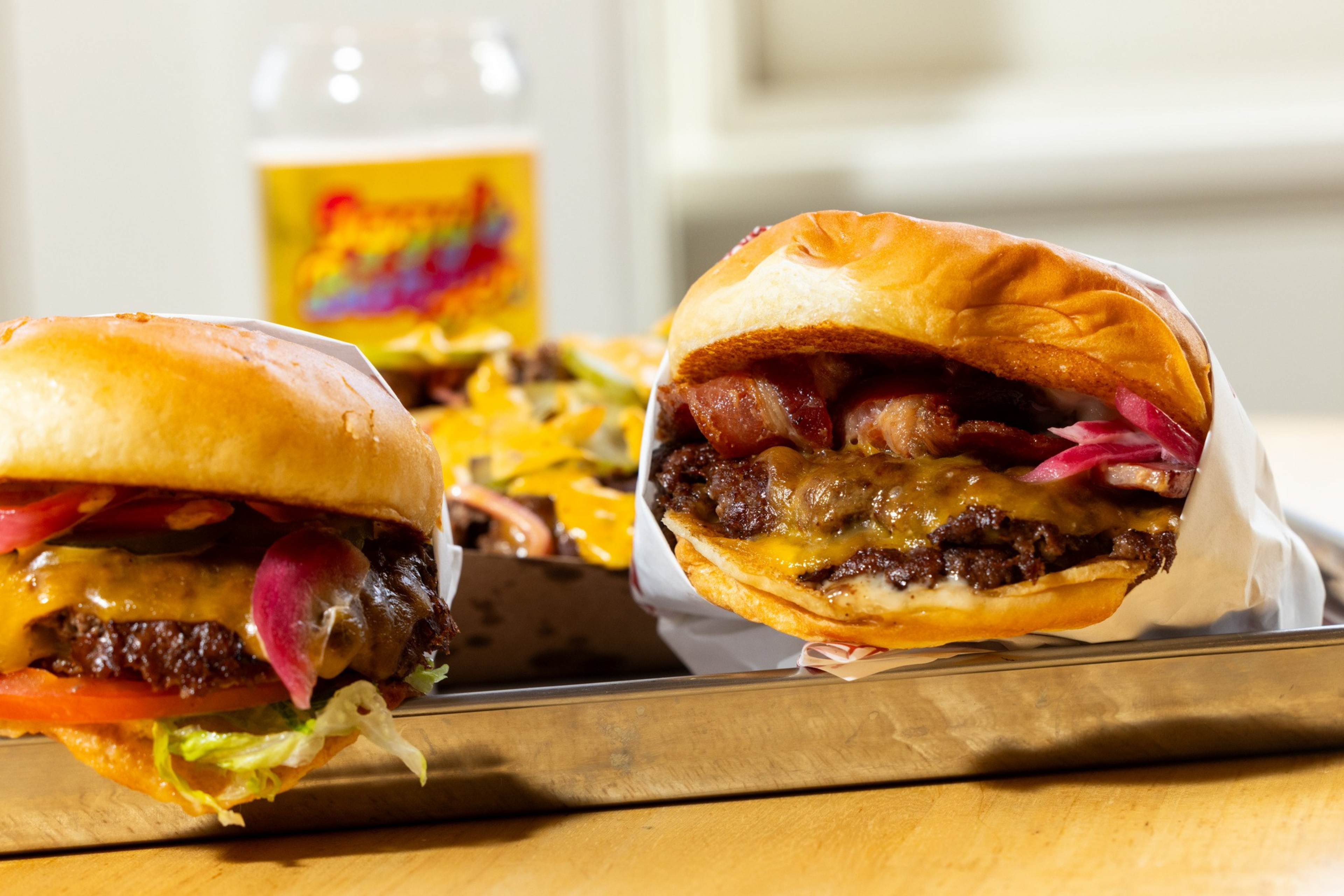 Two cheeseburgers with lettuce, tomato, pickled onions, and bacon rest on a metal tray, with nachos and a beer glass blurred in the background.