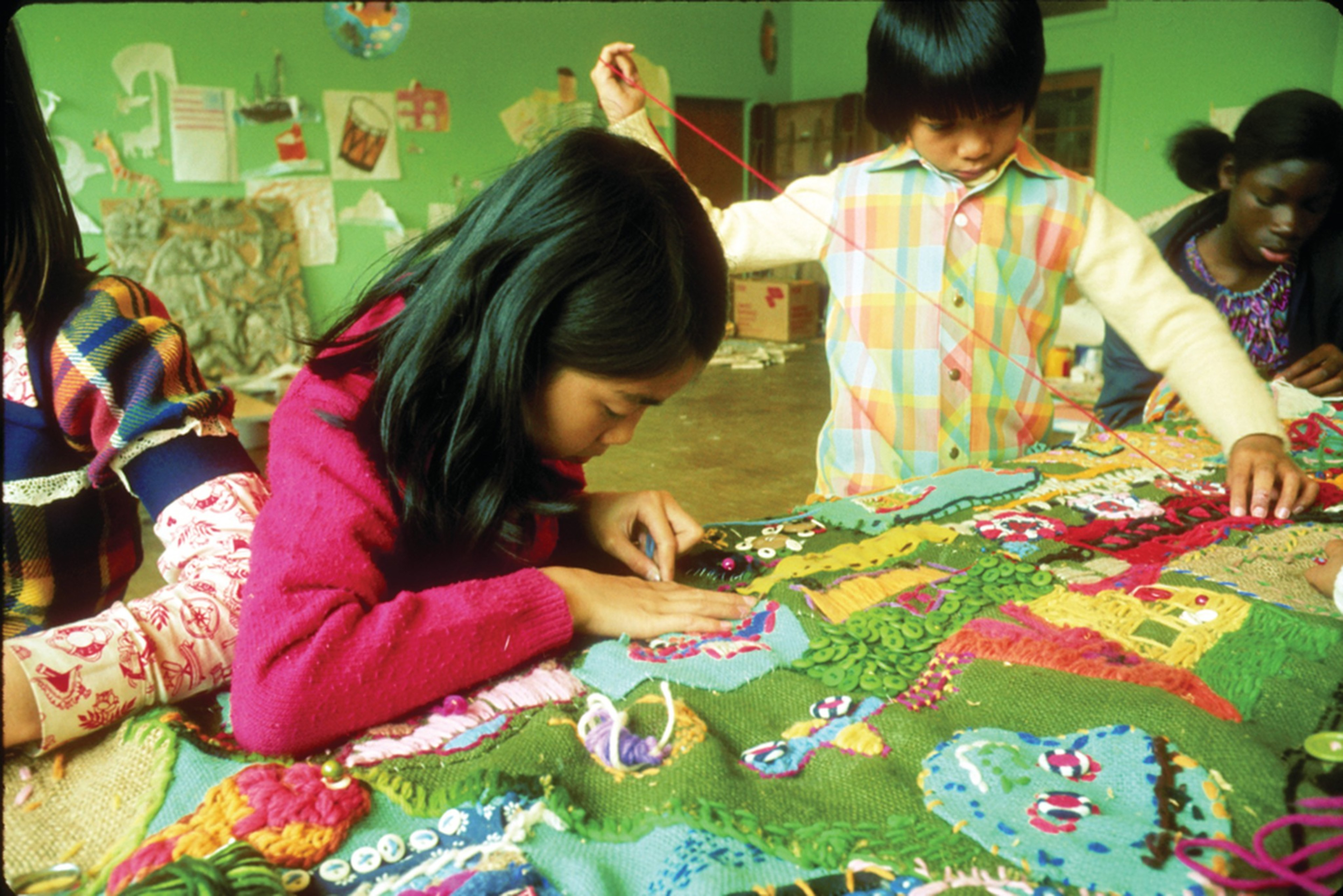 Children are focused on a colorful, detailed textile project, featuring buttons and yarn. The background displays drawings on a green wall.