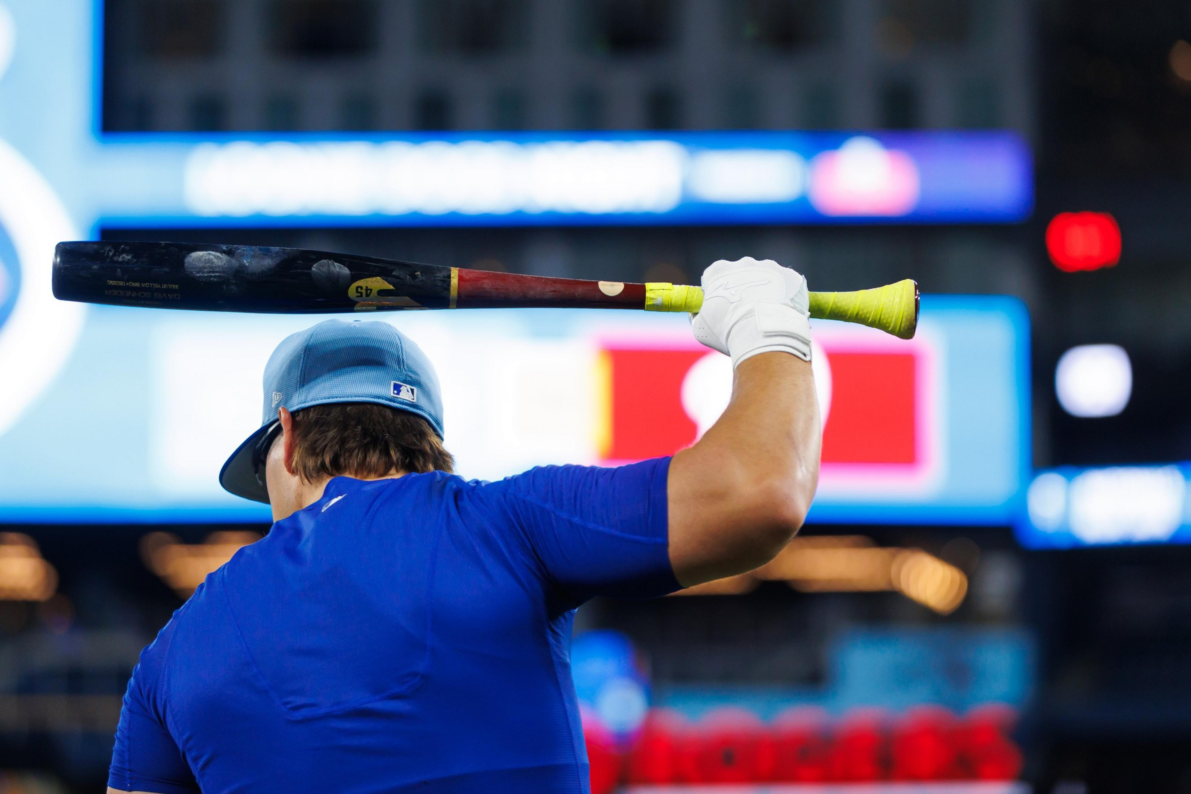 A baseball player in a blue jersey and cap holds a bat over their shoulder, with a blurred stadium and scoreboard in the background.