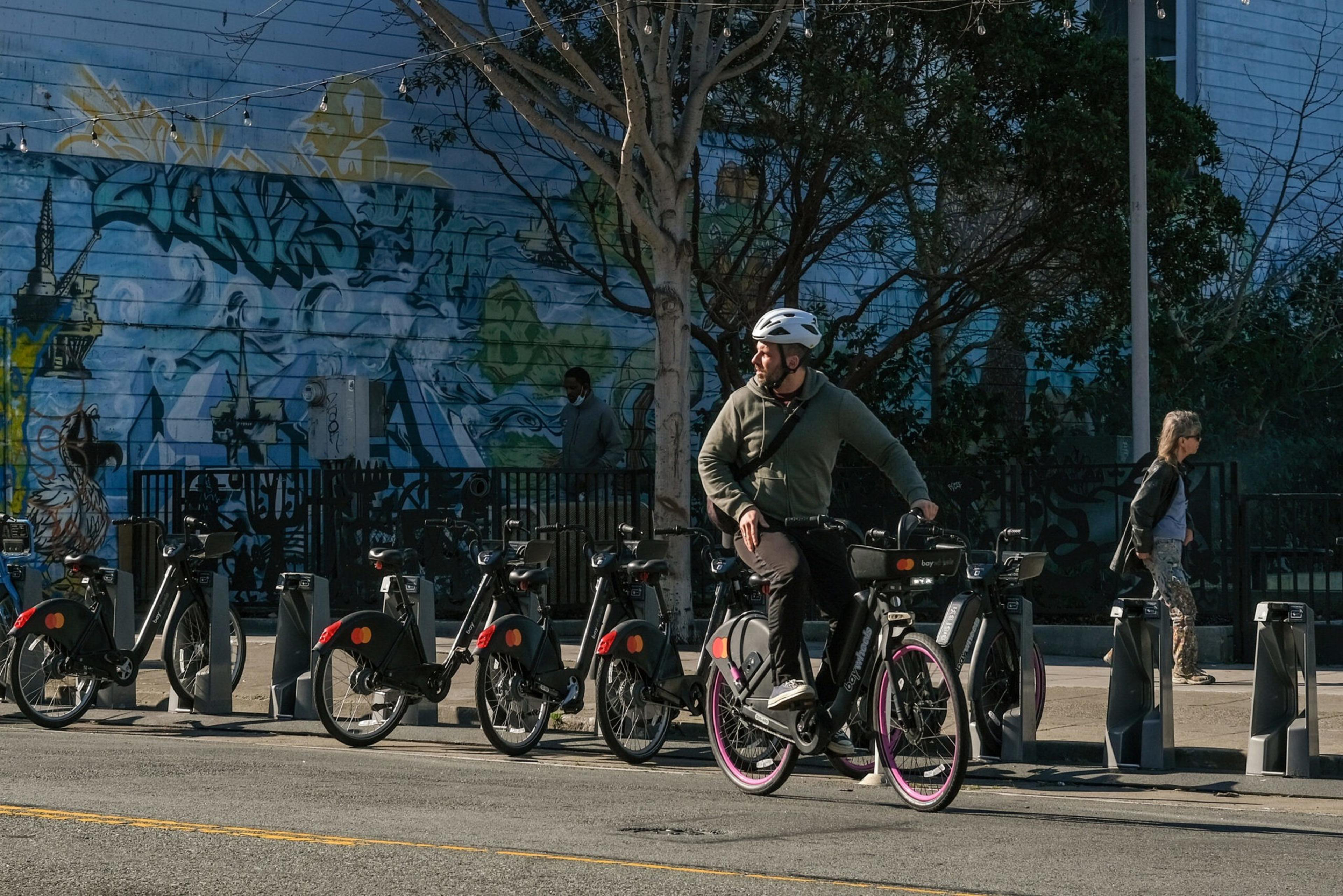 A person wearing a helmet rides a black bike from a row of parked bikes near a graffiti-covered blue wall, while two people walk on the sidewalk.