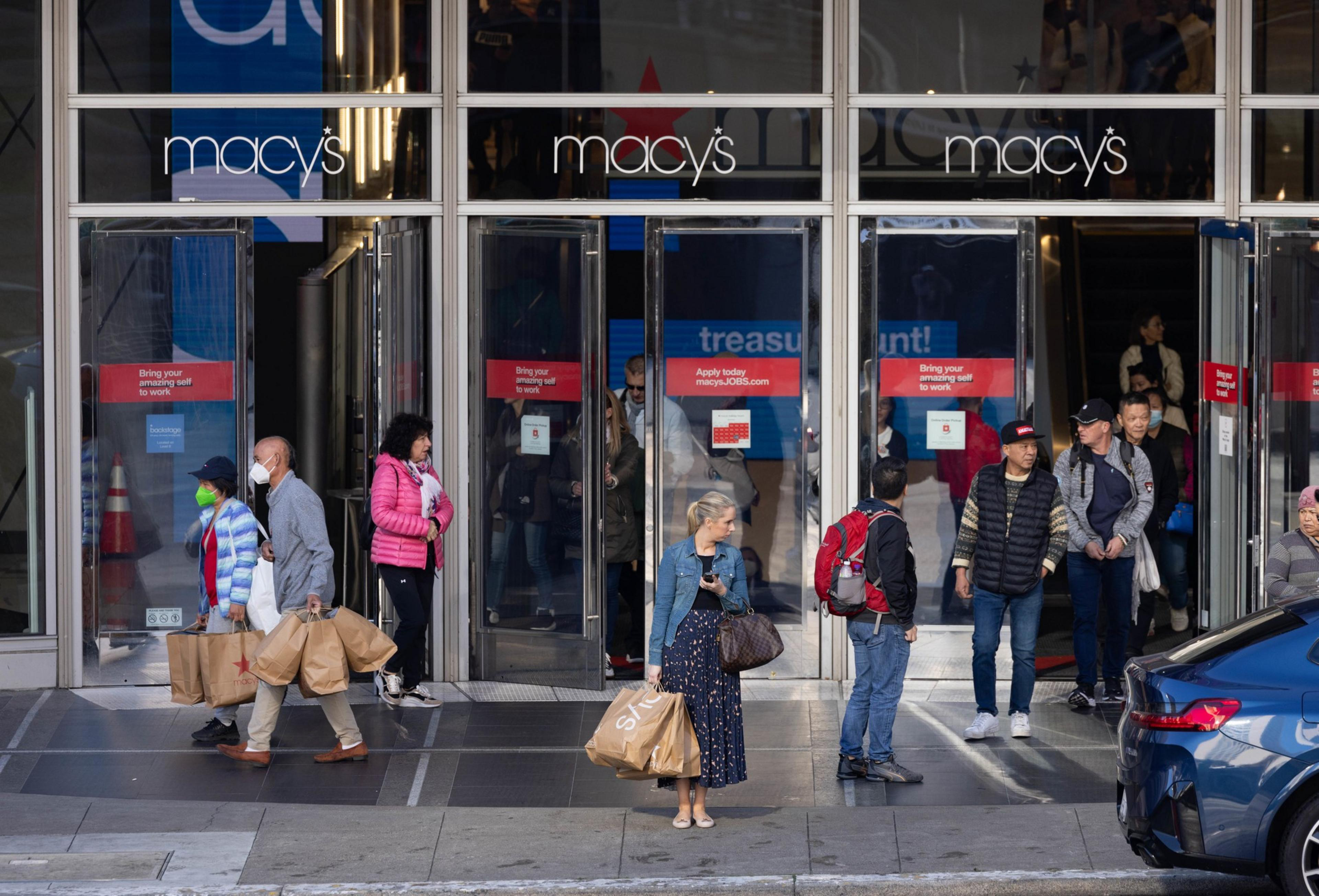 People are exiting a Macy's store carrying shopping bags. Some are wearing masks, and the scene is bustling with a mix of individuals near the shop's entrance.