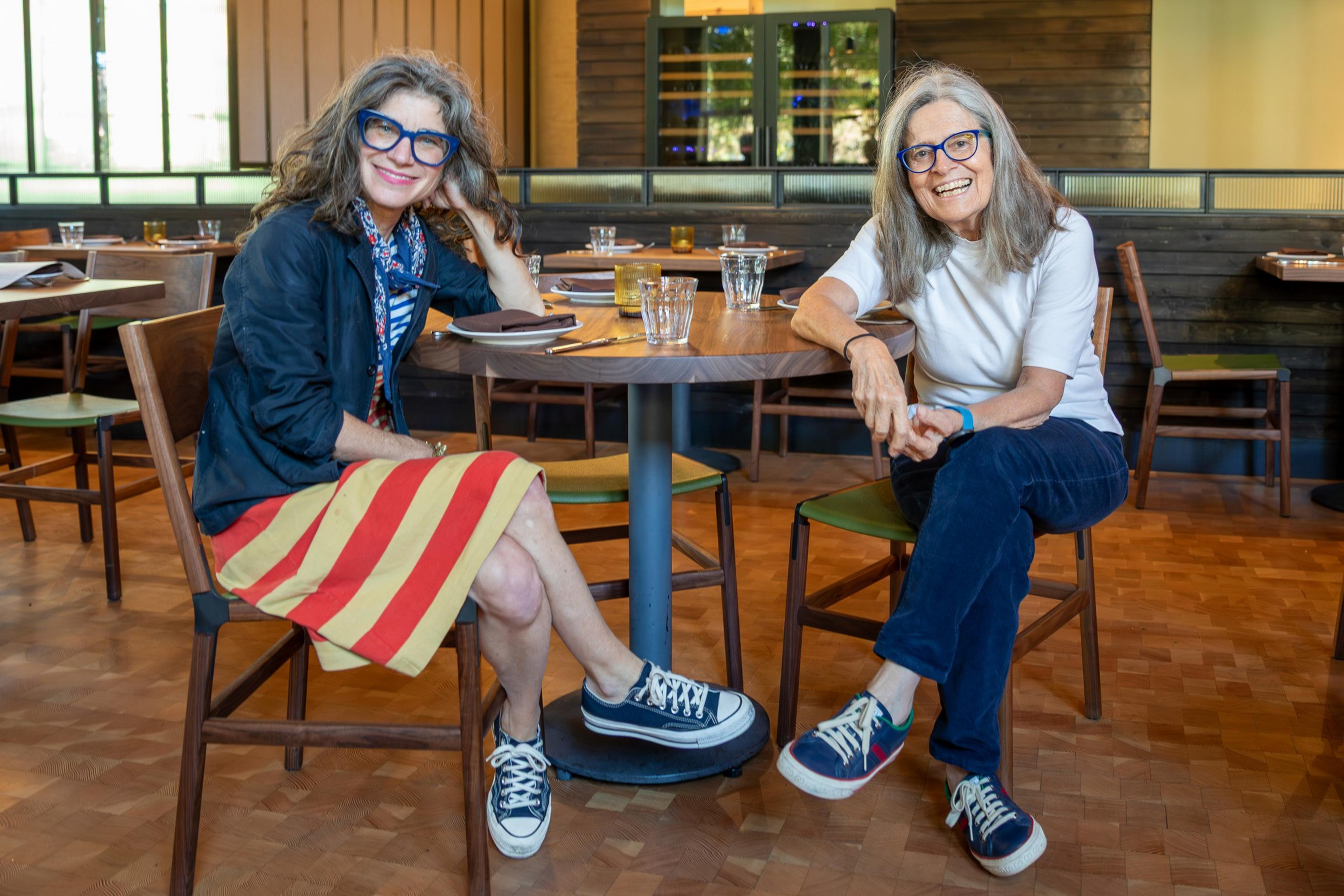 Two women with long hair and blue glasses sit casually at a wooden table in a restaurant. One wears a striped skirt and jacket, the other a white shirt and jeans.