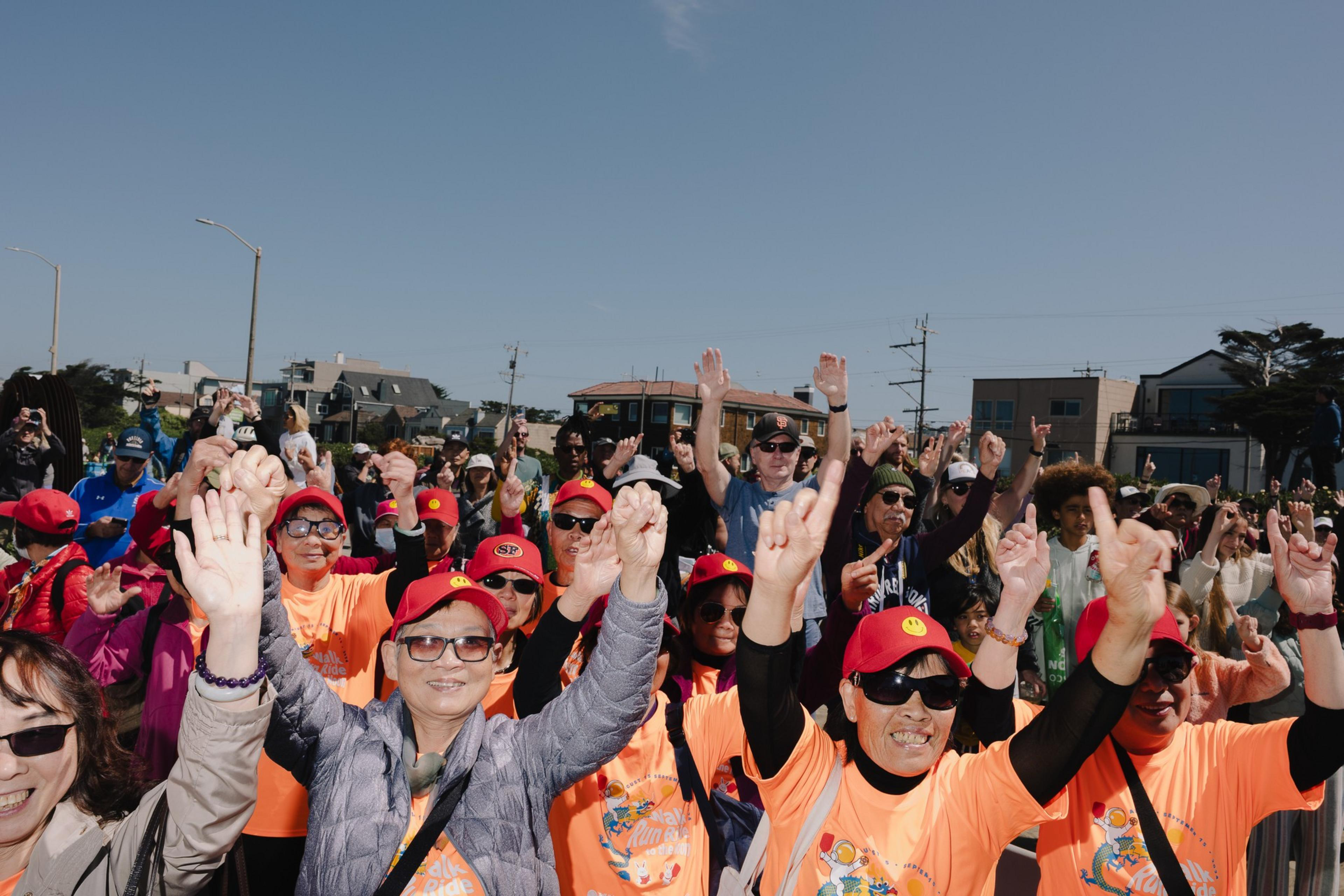 A group of people, wearing orange shirts and red hats, joyfully raise their hands in celebration under a clear blue sky, with buildings in the background.