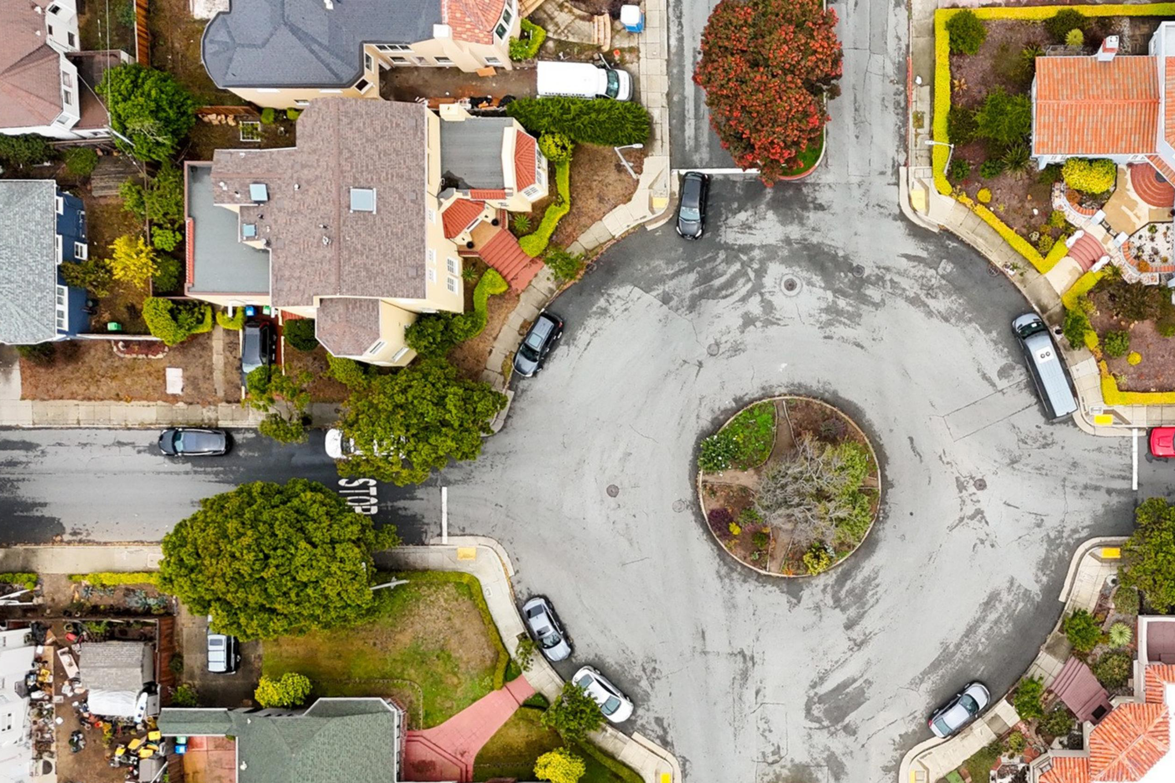 A roundabout intersection with cars parked along the streets and houses surrounding it with trees and gardens nearby.