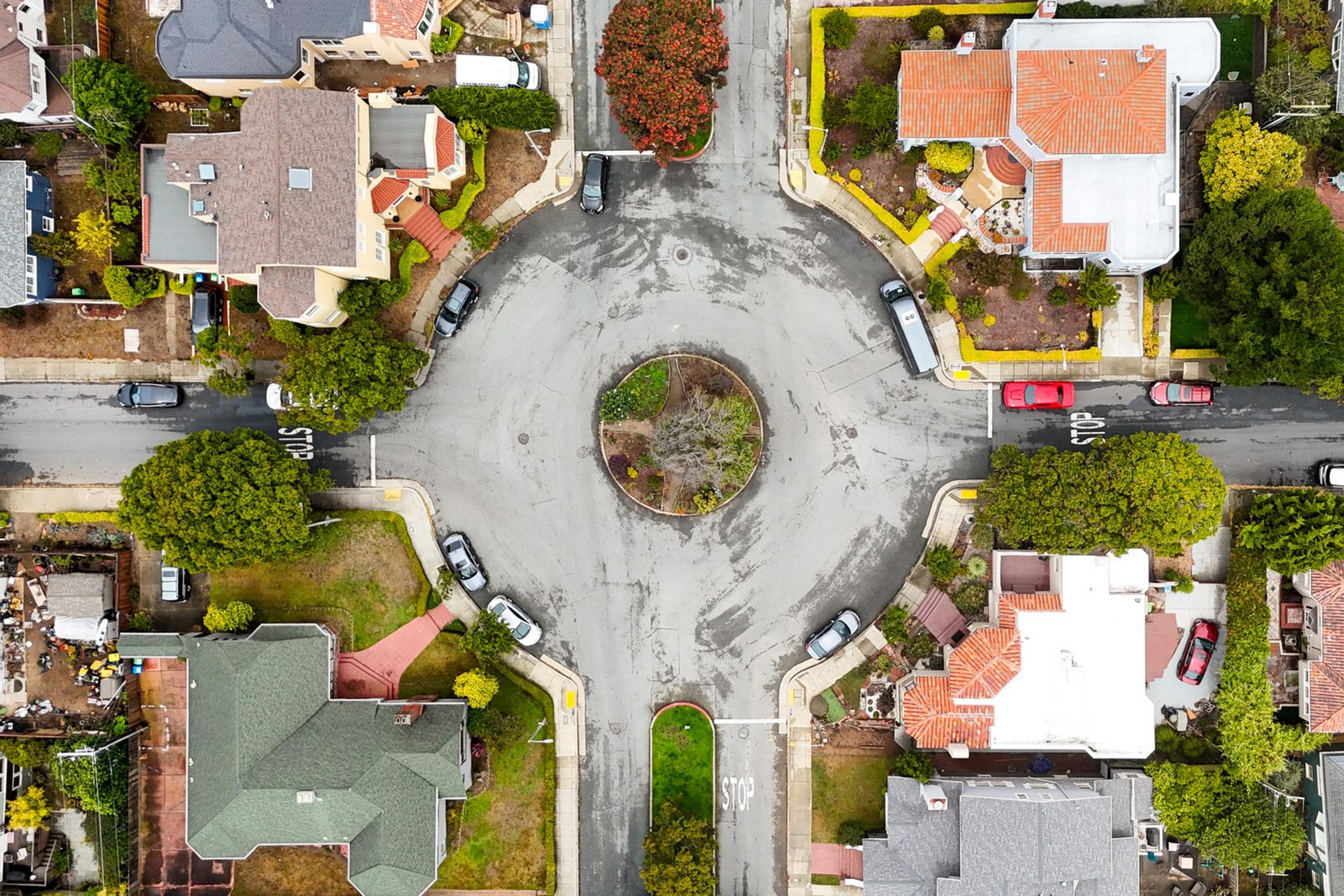 A roundabout intersection with cars parked along the streets and houses surrounding it with trees and gardens nearby.