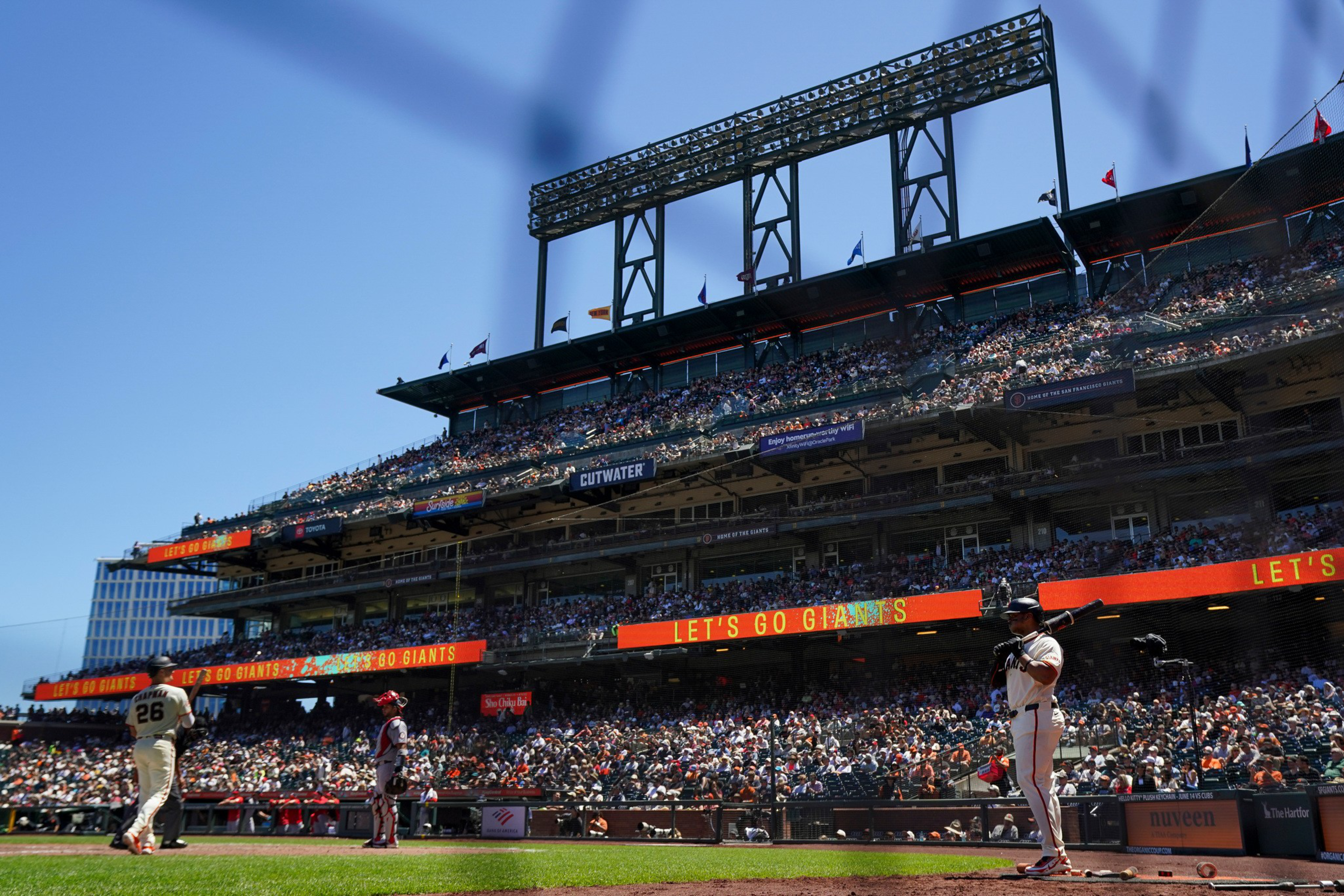 A baseball player in a white uniform bats while another player in a white uniform stands on the field near home plate at a sunny stadium filled with spectators.