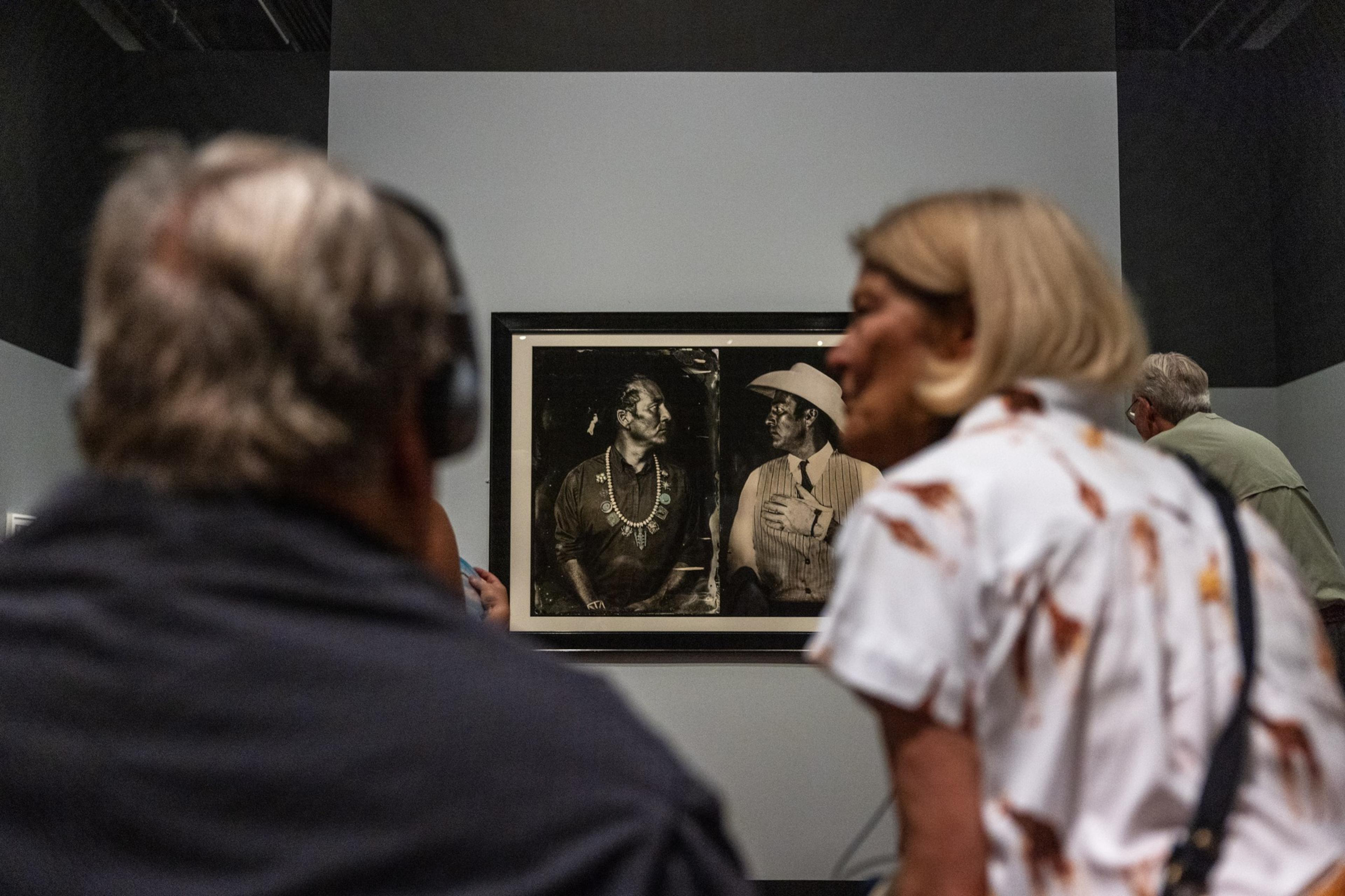 Two people are looking at a black-and-white framed photo of two Native American men facing each other, one wearing a necklace and the other a cowboy hat.