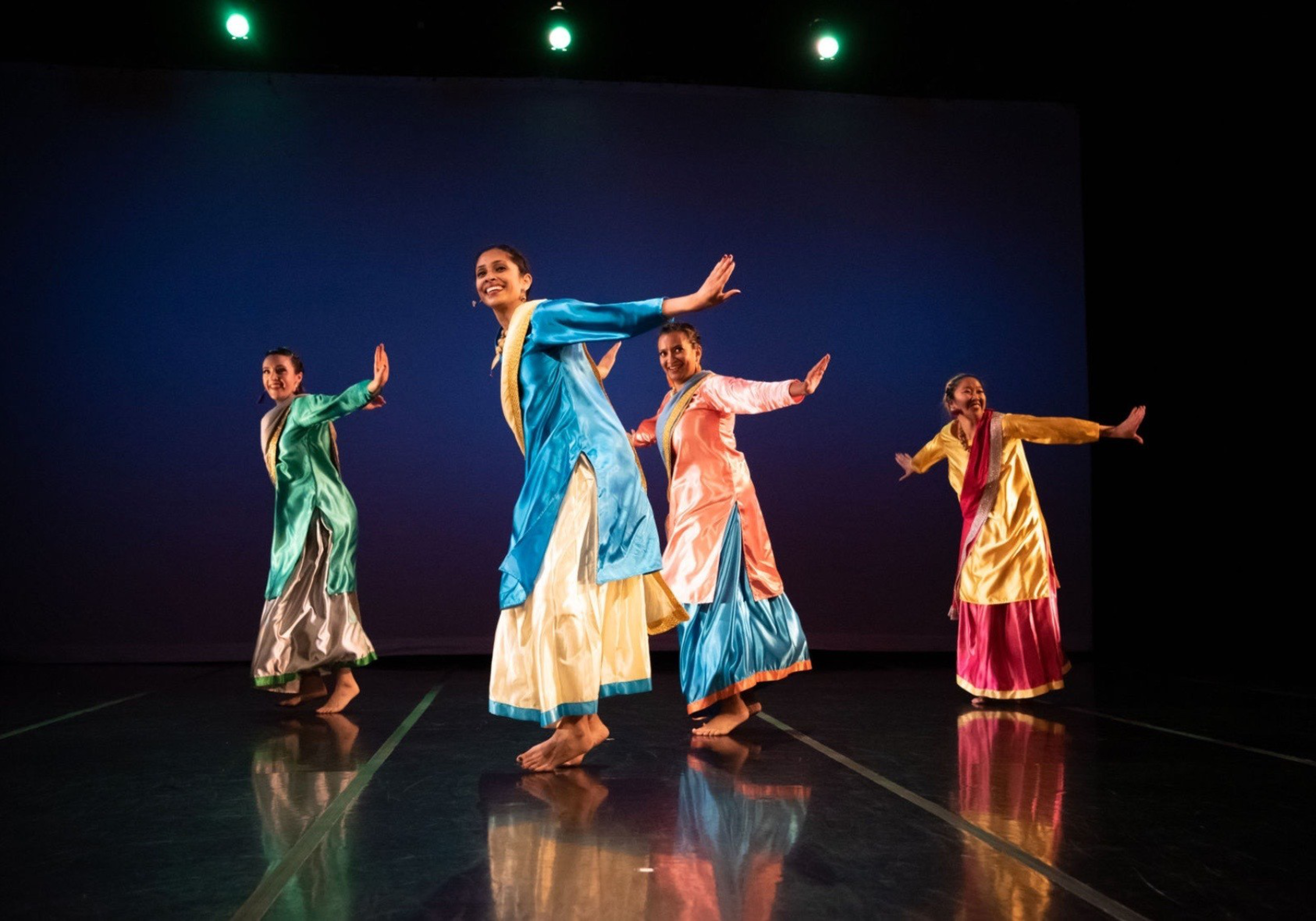 Four women dressed in colorful traditional Indian attire perform a synchronized dance on a reflective stage with a dark blue background.