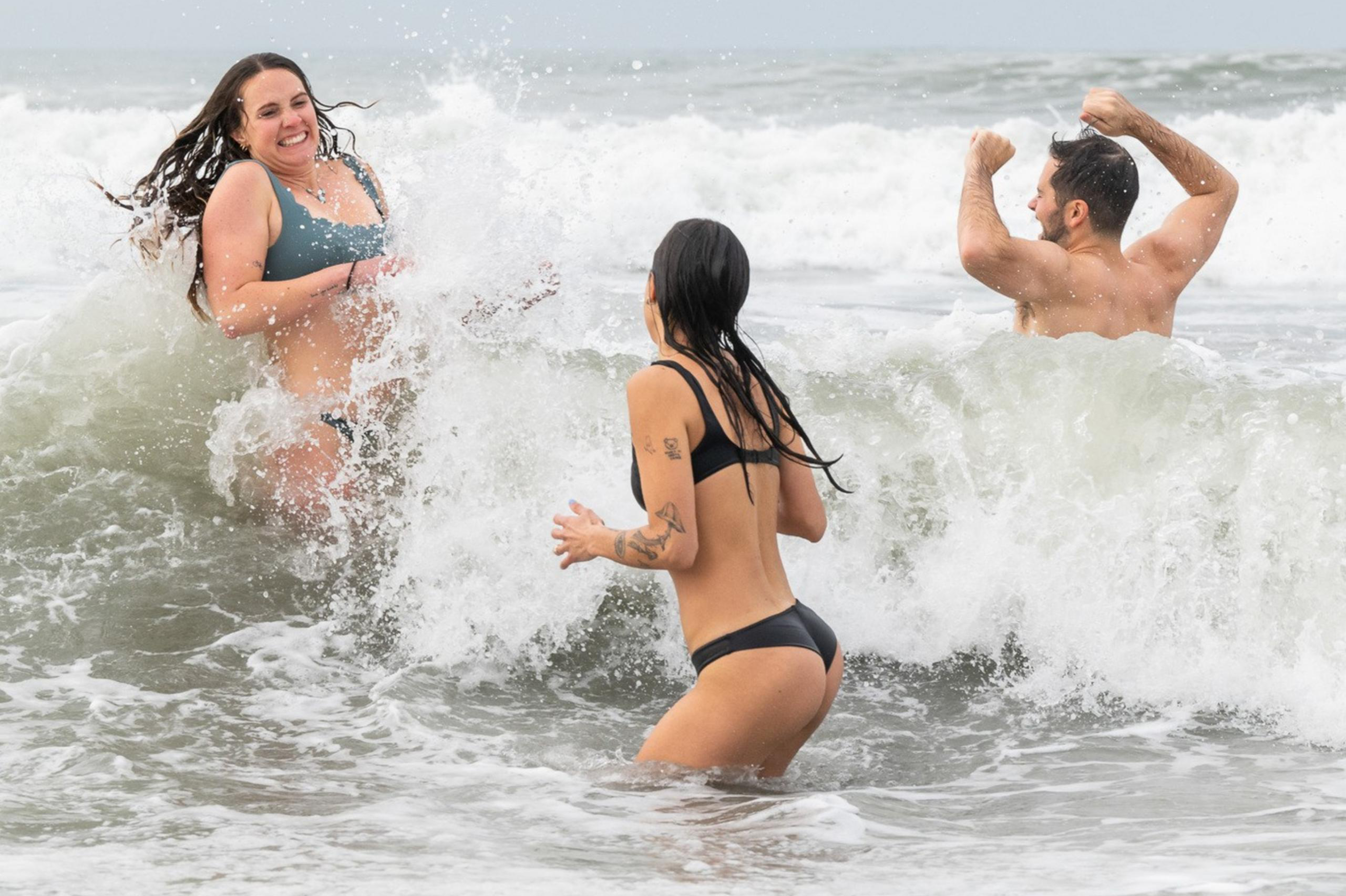 Three people joyfully play in ocean waves, two women in bikinis and a man in swim trunks, splashing and laughing under a cloudy sky.