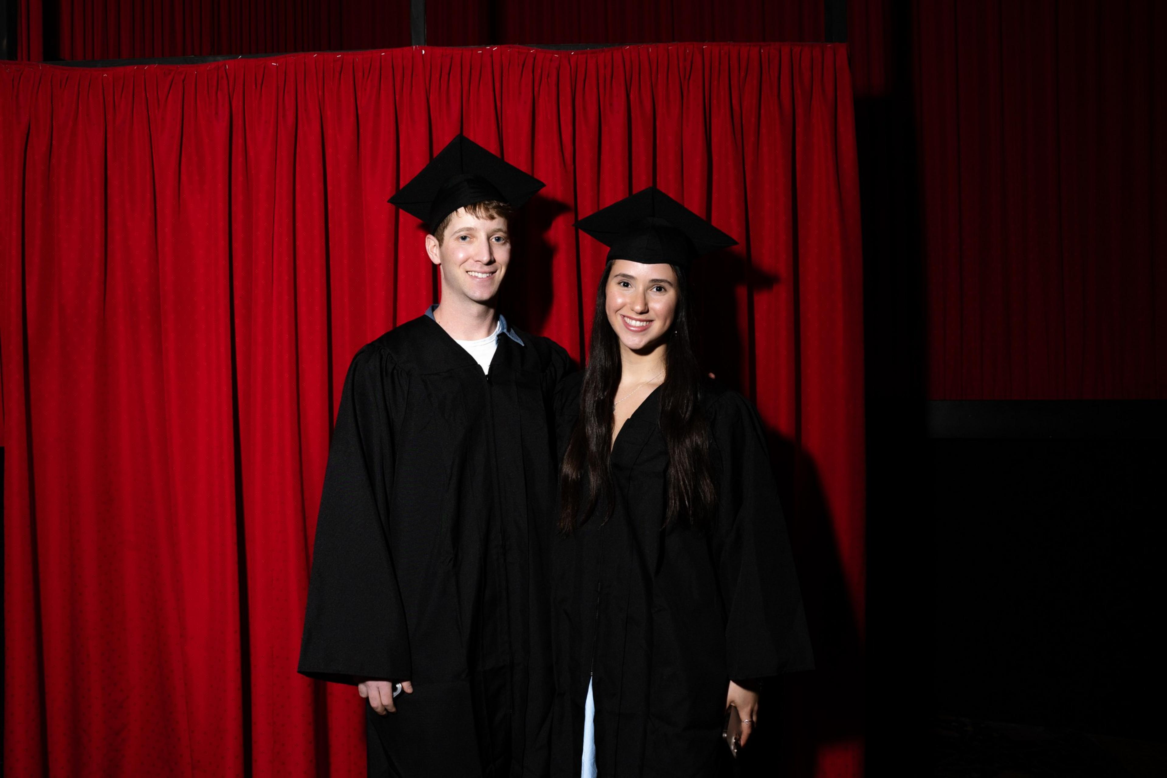Two people in graduation gowns and caps stand smiling in front of a red curtain backdrop.