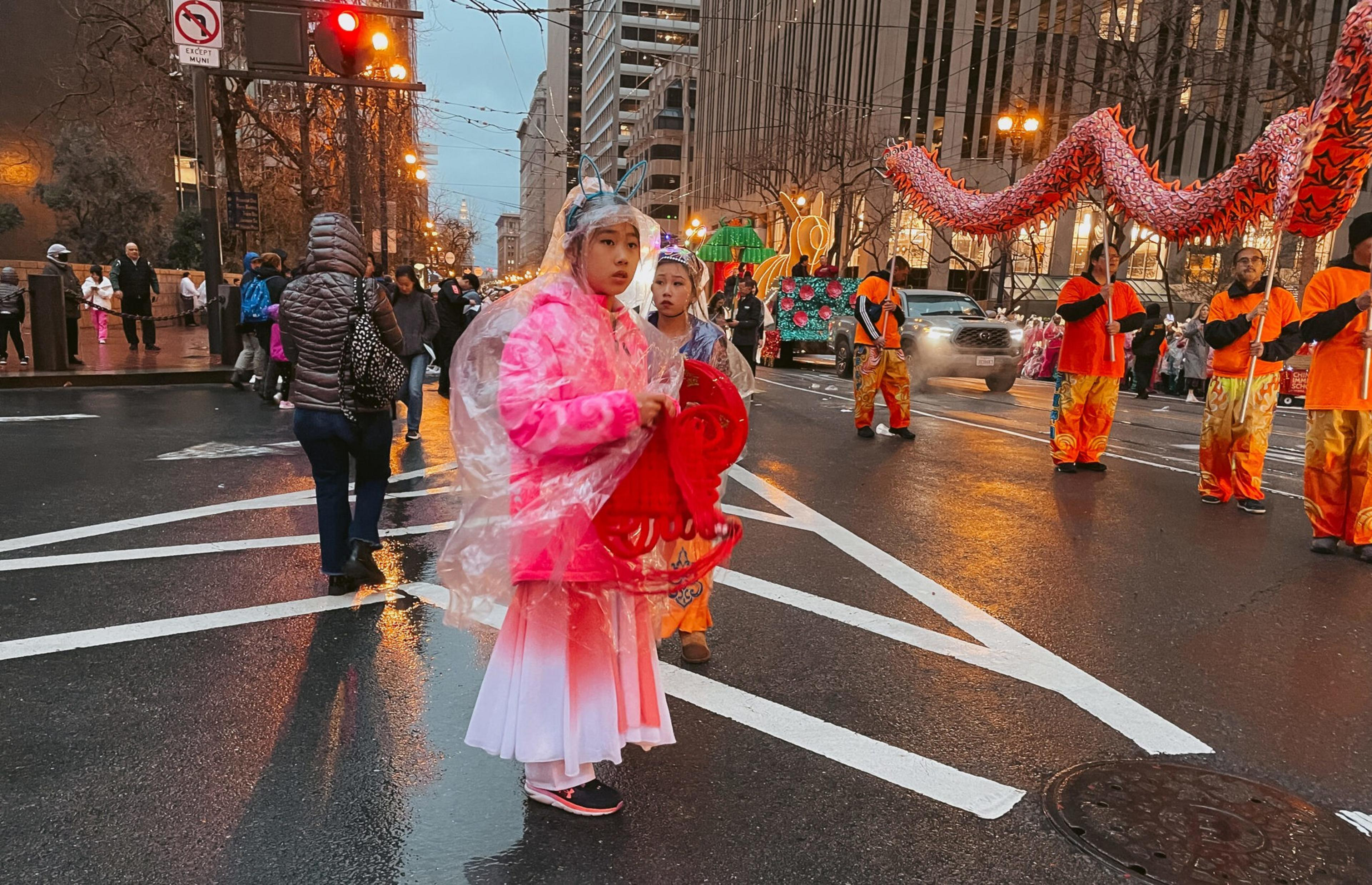 Children in festive costumes and rain ponchos stand on a wet city street during a parade featuring performers holding a red dragon.