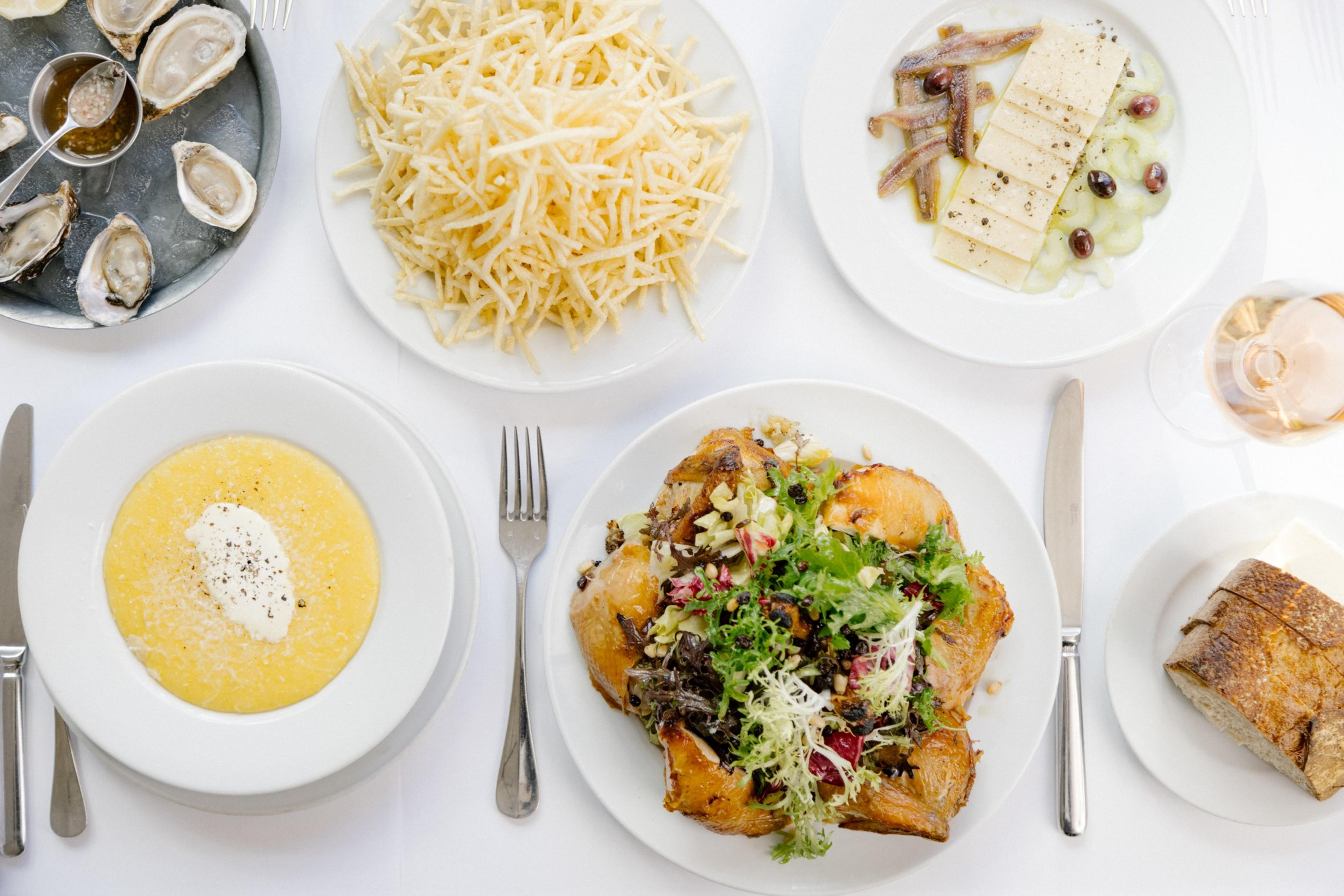 An overhead view of bowl of polenta with mascarpone, oysters, shoestring potatoes, house-cured anchovies with celery, Parmigiano-Reggiano and coquillo olives, bread, and chicken for two roasted in the wood-fired oven served with a warm bread salad with scallions, garlic, bitter greens, dried currant and pine nuts at Zuni Café .
