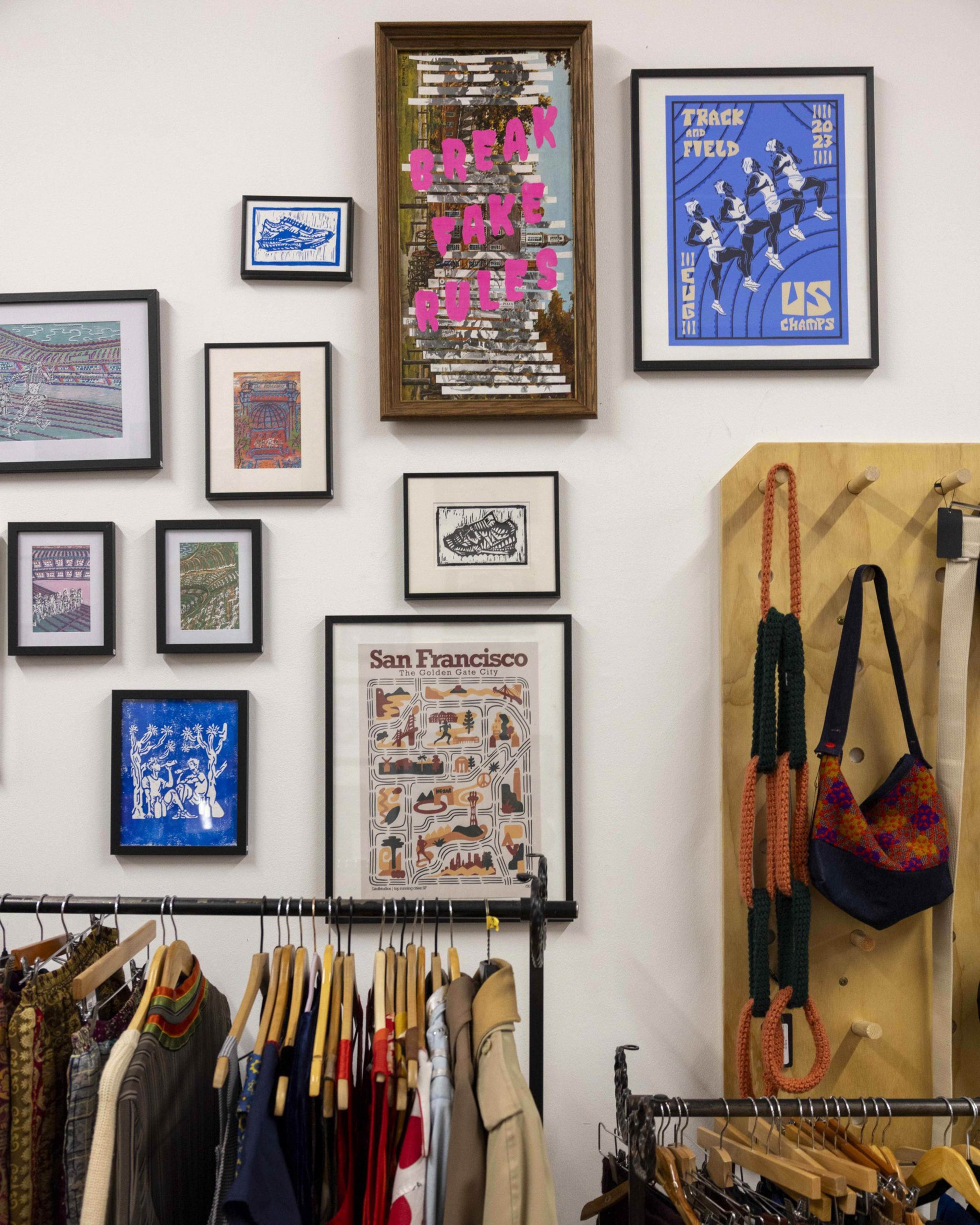 A wall with framed art and prints, including one saying &quot;Break Fake Rules.&quot; Below, a rack holds colorful clothes, and to the right, a purse hangs on pegs.