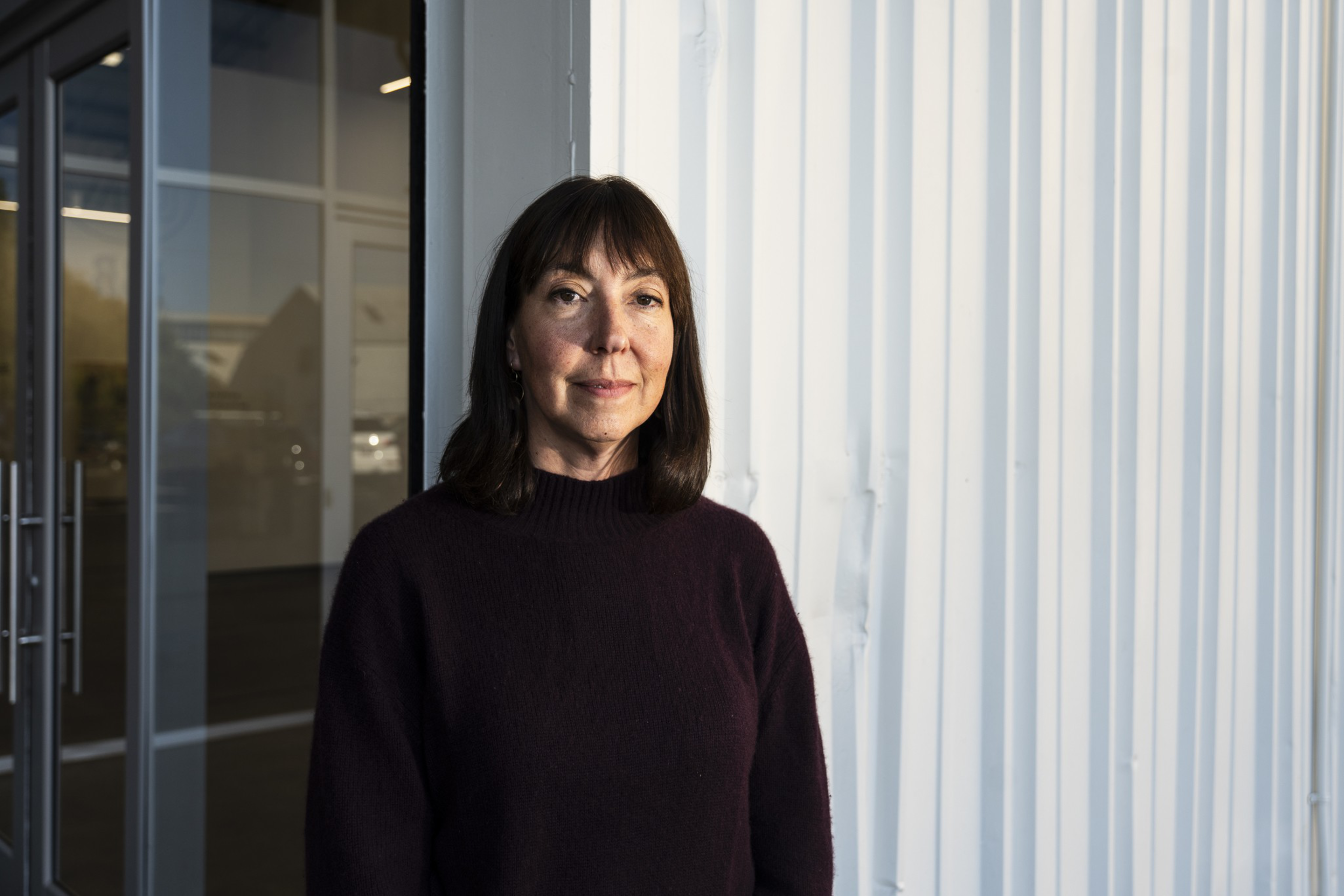 A woman with shoulder-length dark hair wears a dark sweater, standing indoors near a glass door and white paneled wall, softly lit from the side.