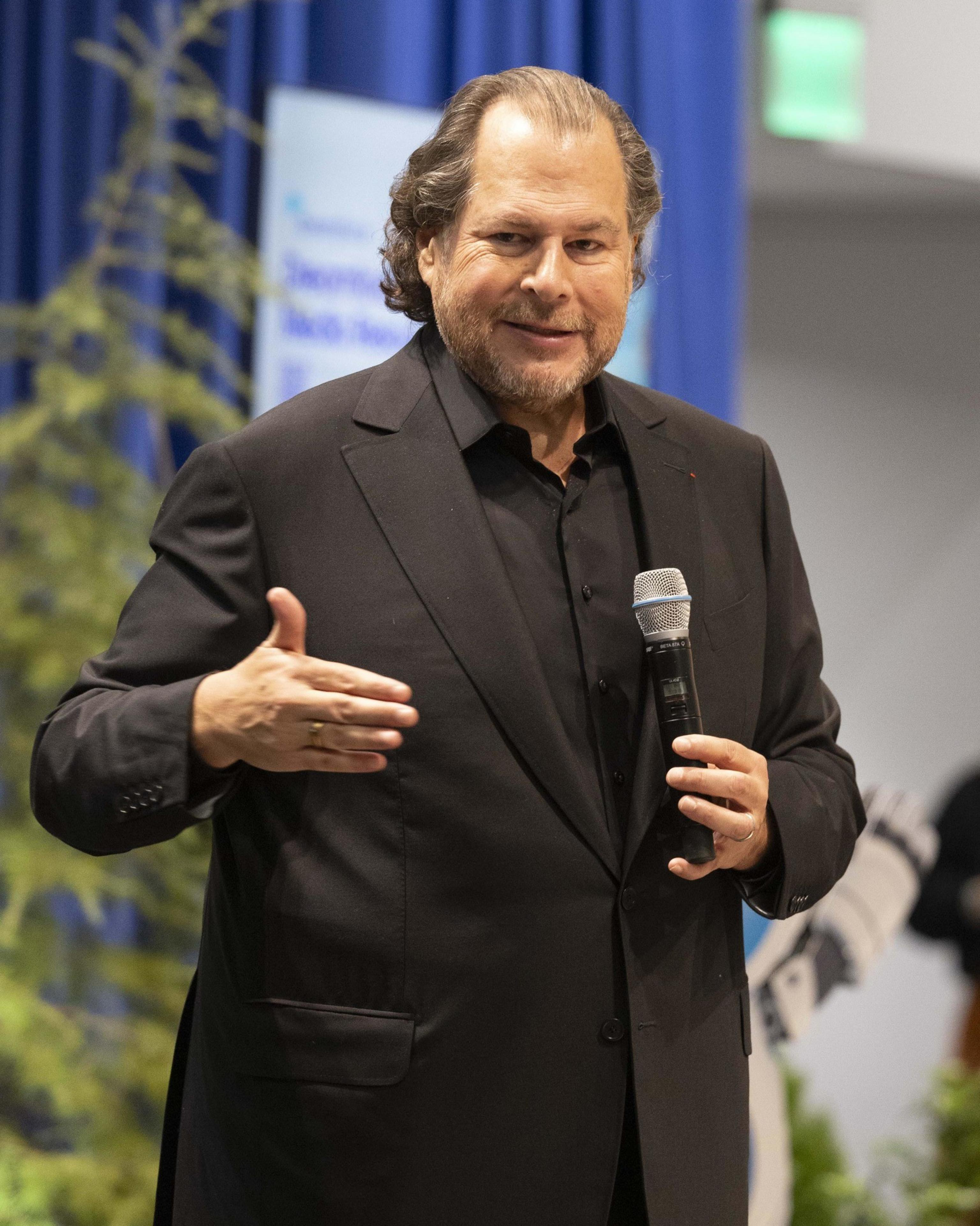 A man in a dark suit and black shirt is holding a microphone while gesturing. He has a short beard and stands in front of a blue curtain with foliage in the background.