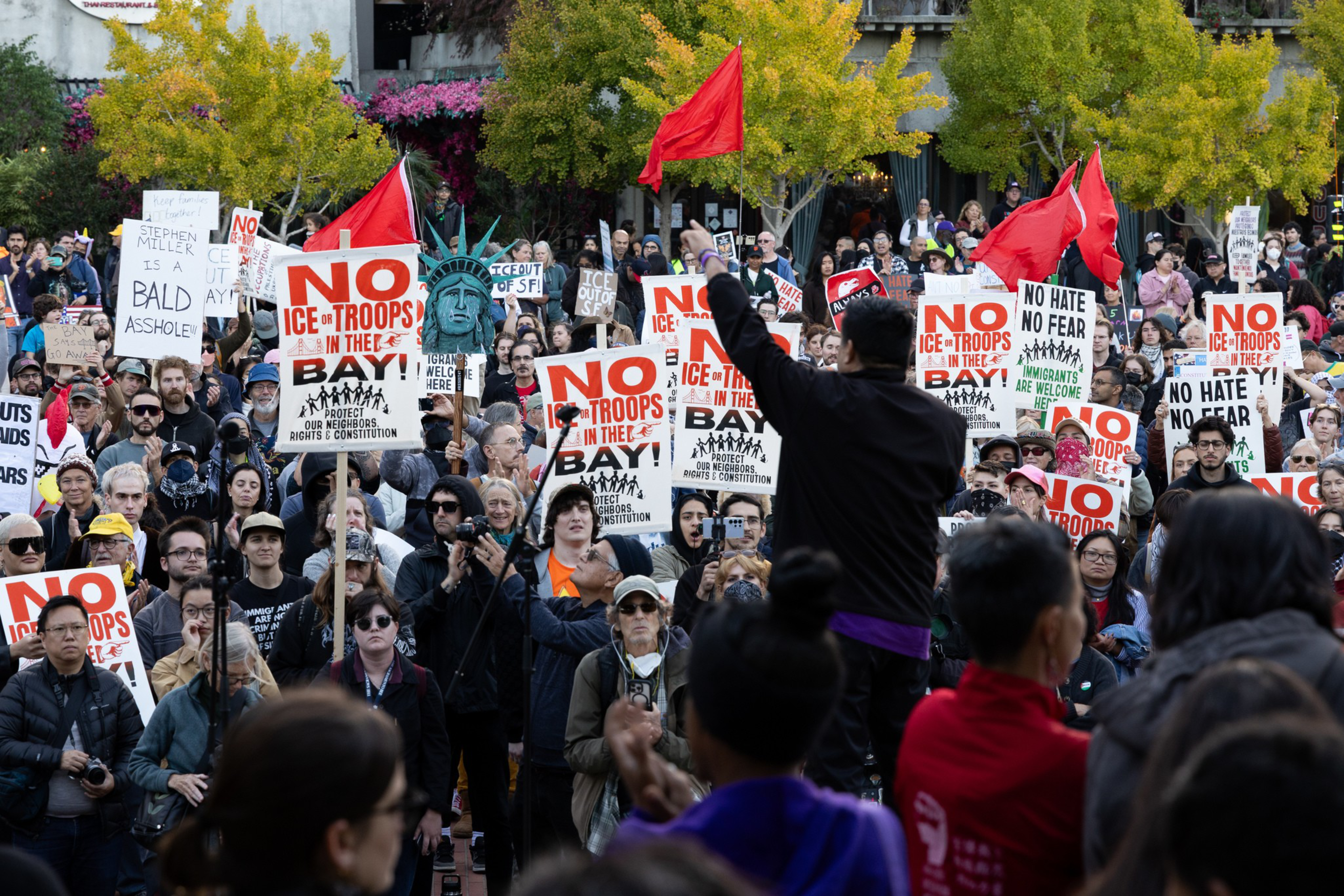 A crowd of protesters holds signs saying “No ICE troops in the Bay” and “No hate no fear,” with red flags and a small Statue of Liberty statue in the background.
