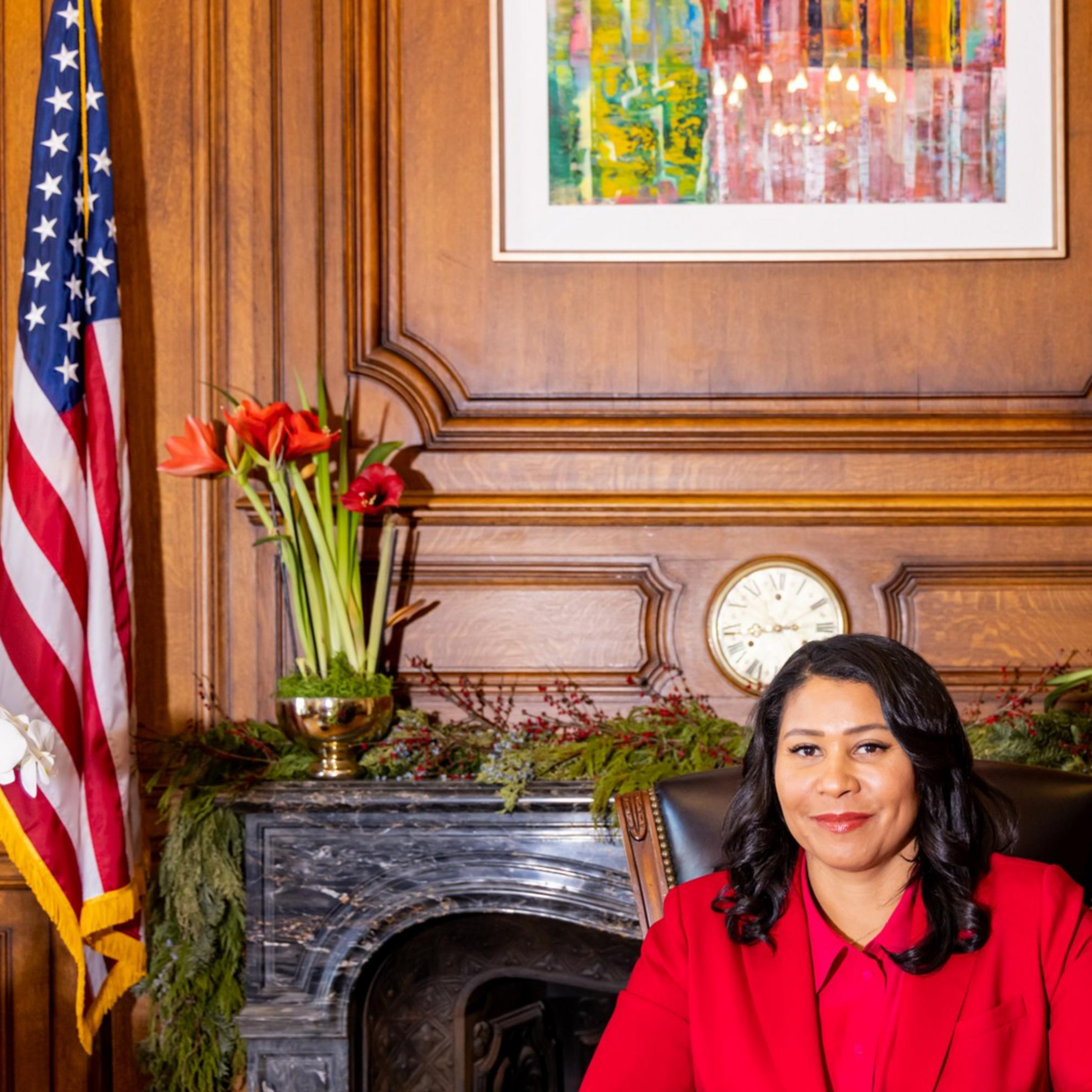 A person in a red outfit sits at a wooden desk in an ornate room. Behind them are flags, a clock, and flowers, with a colorful painting on the wall.