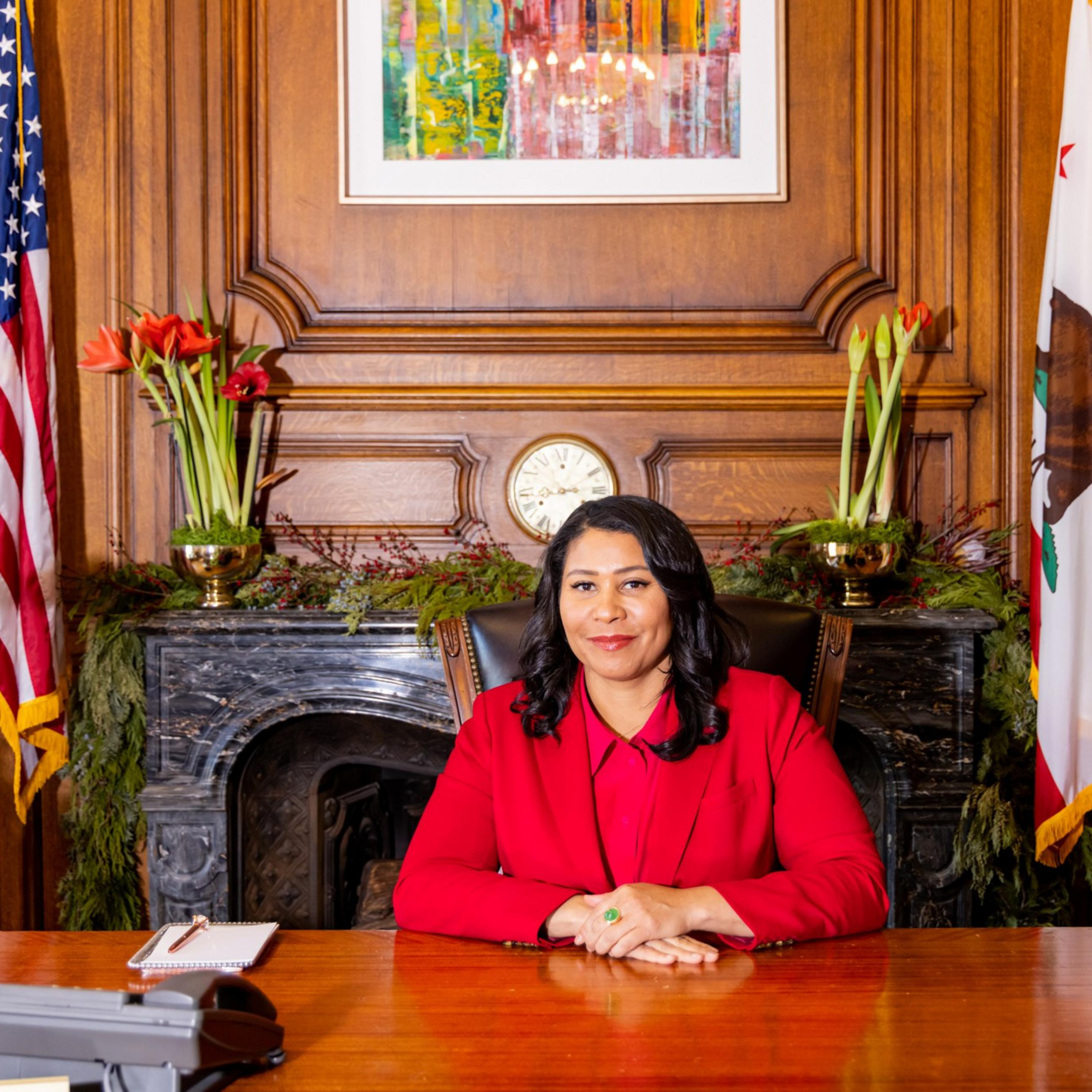 A person in a red outfit sits at a wooden desk in an ornate room. Behind them are flags, a clock, and flowers, with a colorful painting on the wall.