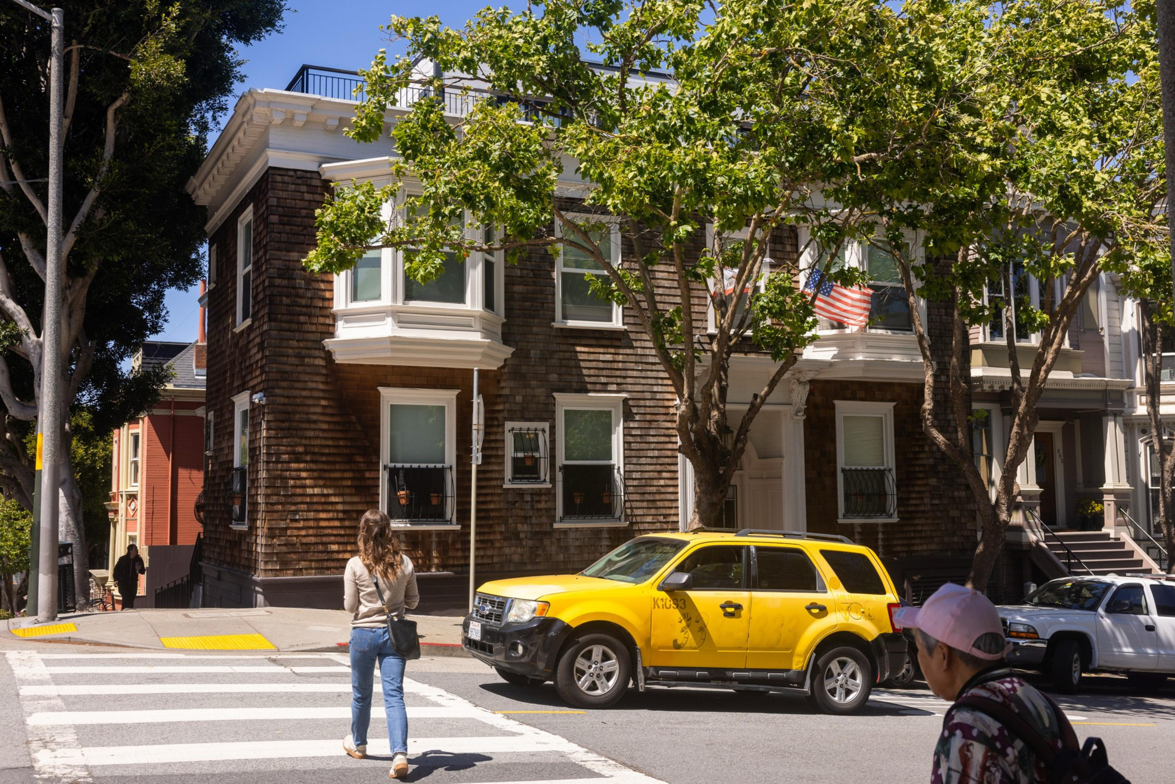 A woman crosses a street towards a corner house with wooden siding and a tree in front. A yellow SUV is parked nearby, and an American flag hangs from the house.